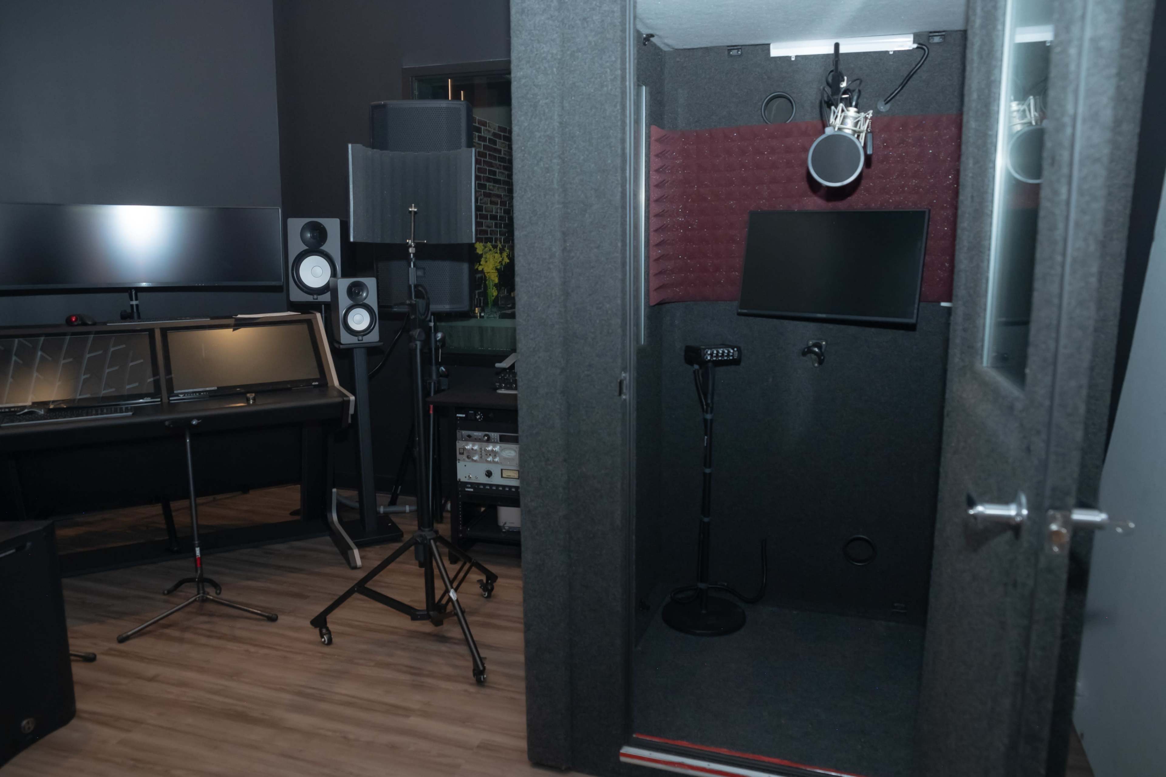 The image shows a sound booth with a microphone, a monitor, and soundproofing panels, set within a recording studio that includes a control desk and speaker setup.