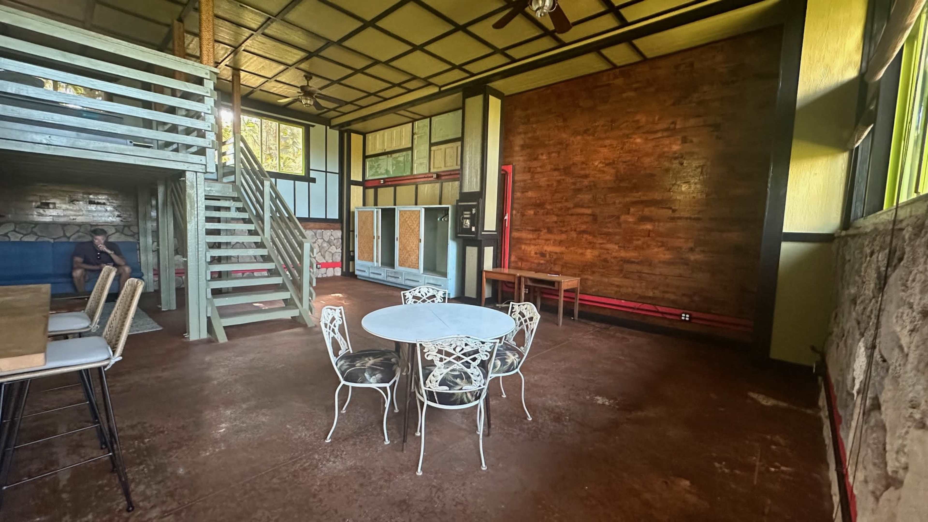 A spacious room features a round dining table with white chairs, a staircase in the background, and a wooden wall accent.