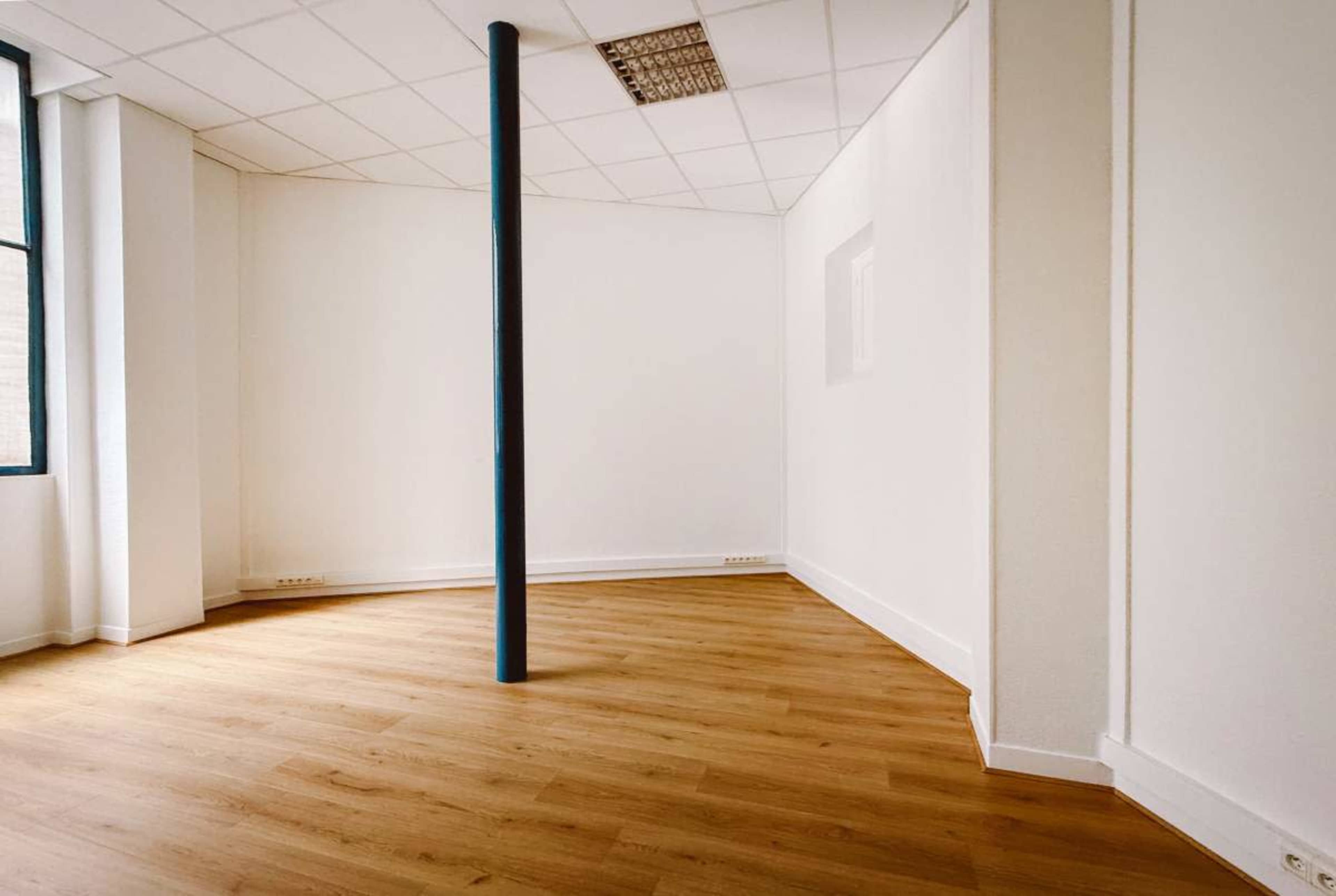 The image shows an empty room with light wood flooring and a single vertical column in the center.