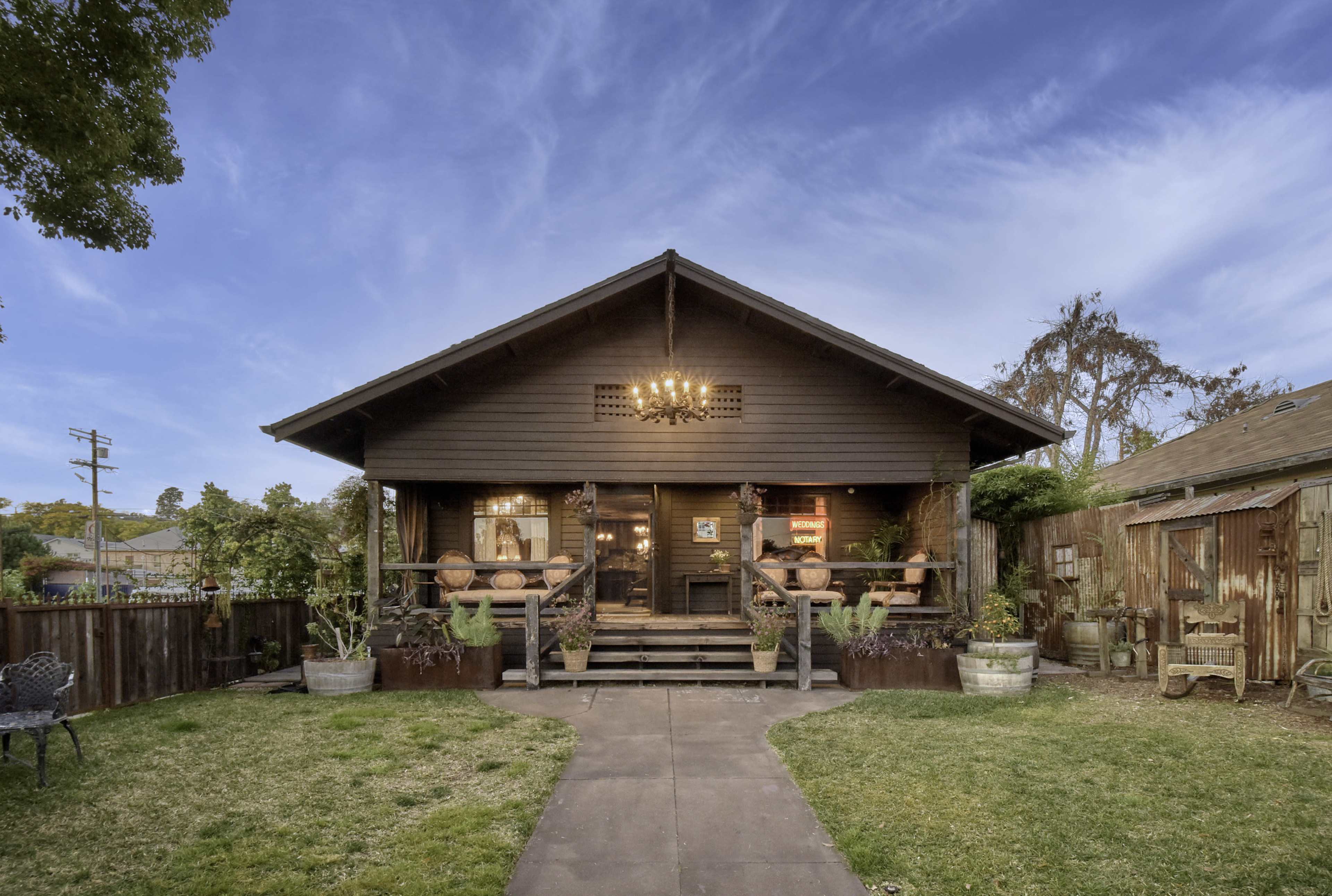A craftsman-style house features a wide front porch with potted plants and a chandelier, set against a clear blue sky.