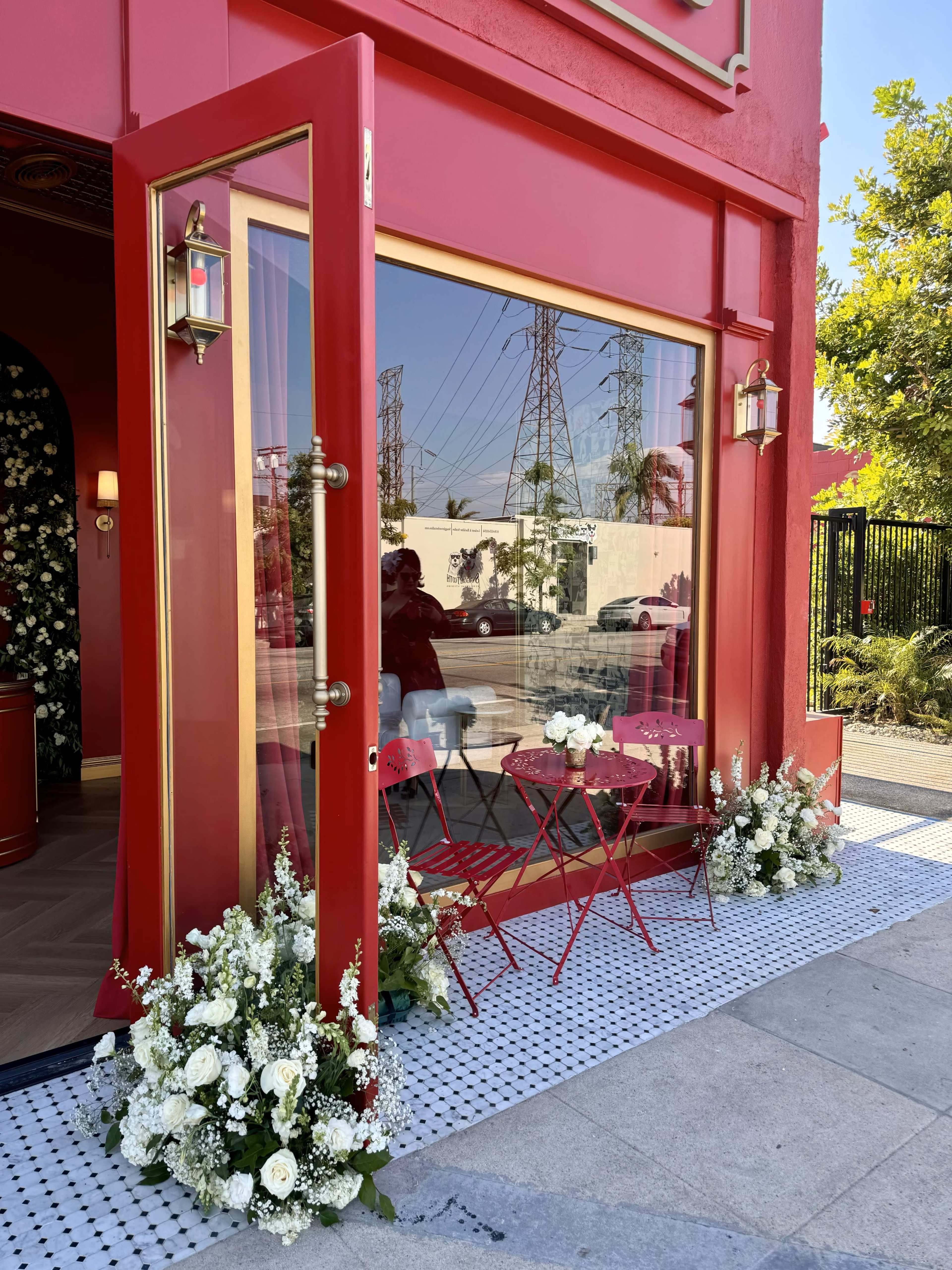 A red storefront features large glass doors, small bistro tables with chairs, and white floral arrangements lining the entrance.