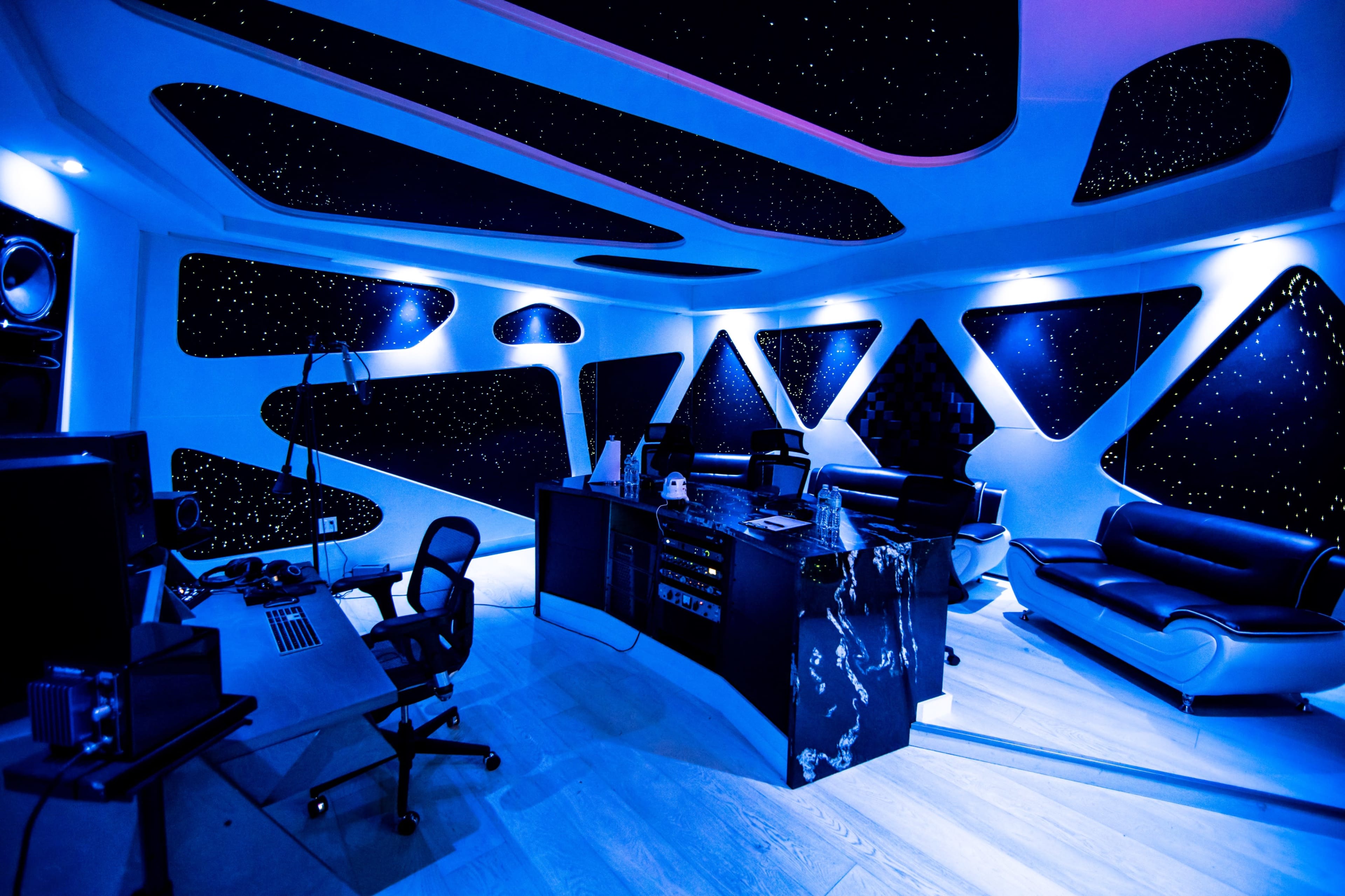 The image shows a modern office space with sleek black furniture, a desk setup with computer equipment, and a ceiling designed to resemble a starry night sky, illuminated by blue lighting.
