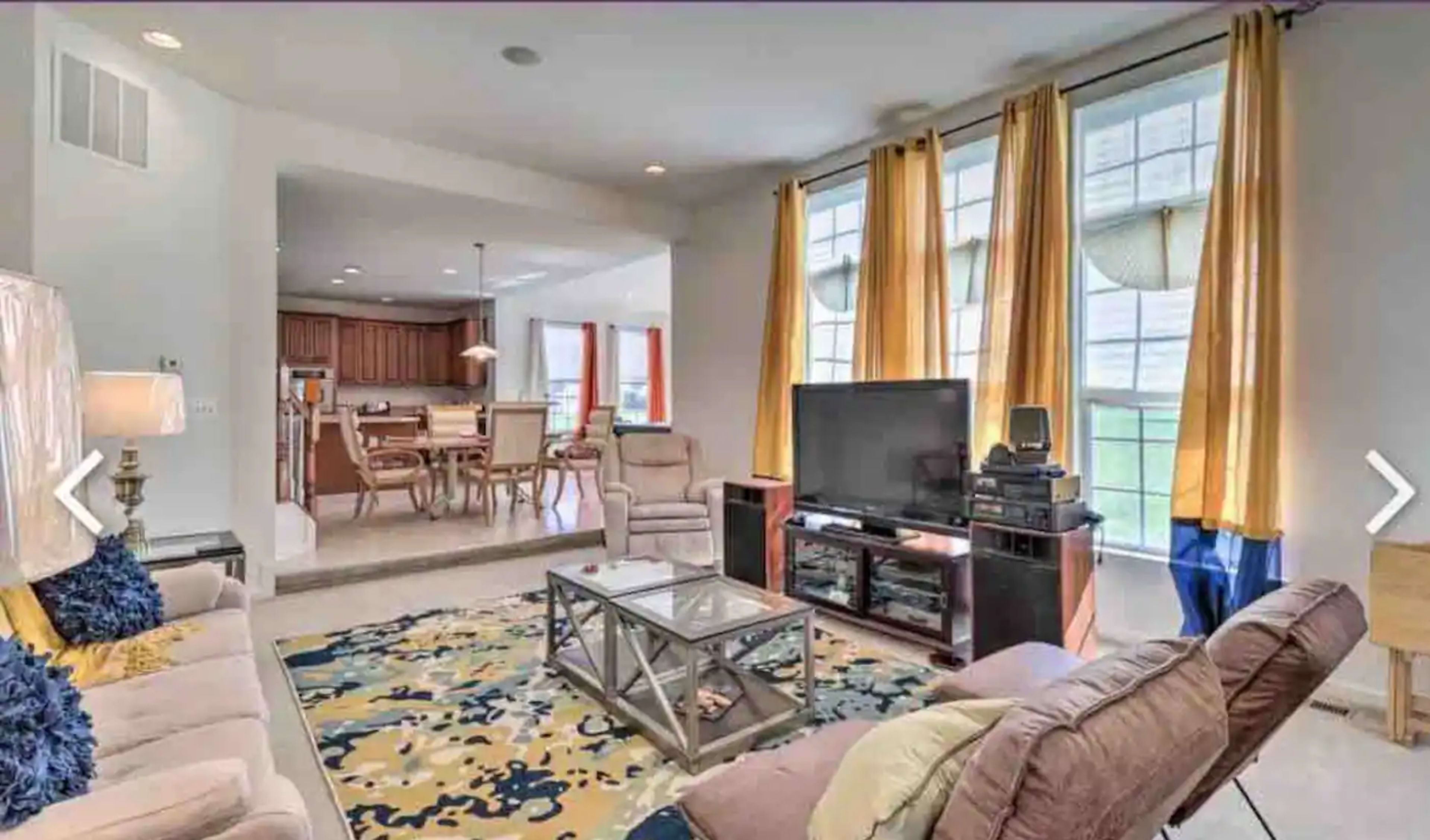 A living room with a large window dressed in yellow curtains, a television on a stand, and a coffee table on a colorful rug, leading into a dining area in the background.