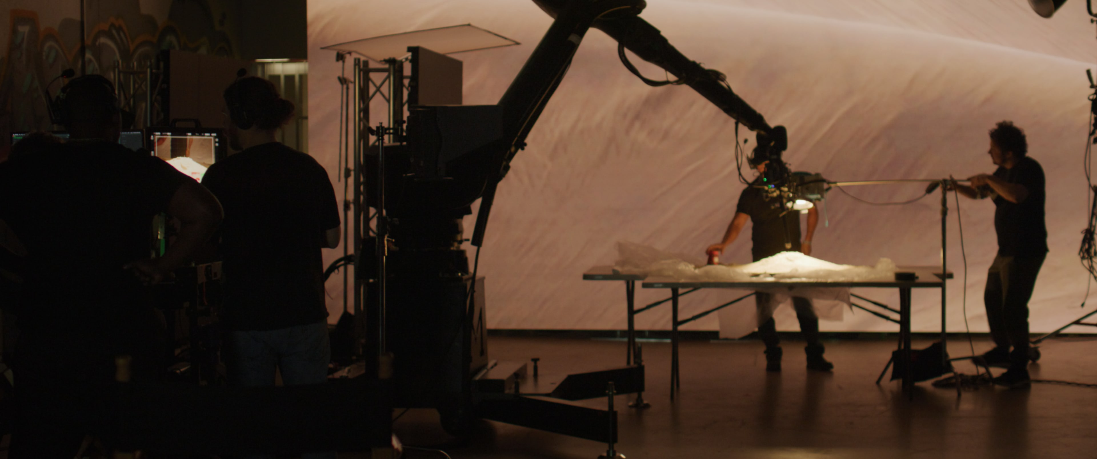 The image shows a film production set with crew members operating equipment around a table lit by bright lights, against a large backdrop.