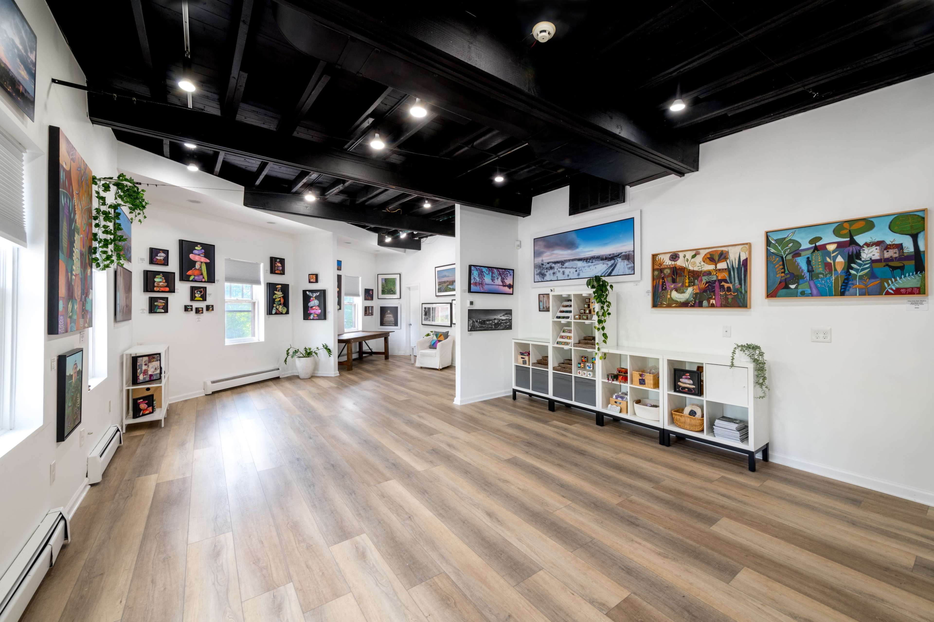 A spacious, modern interior room with wooden flooring, white walls adorned with various artworks, and a minimalistic shelving unit.