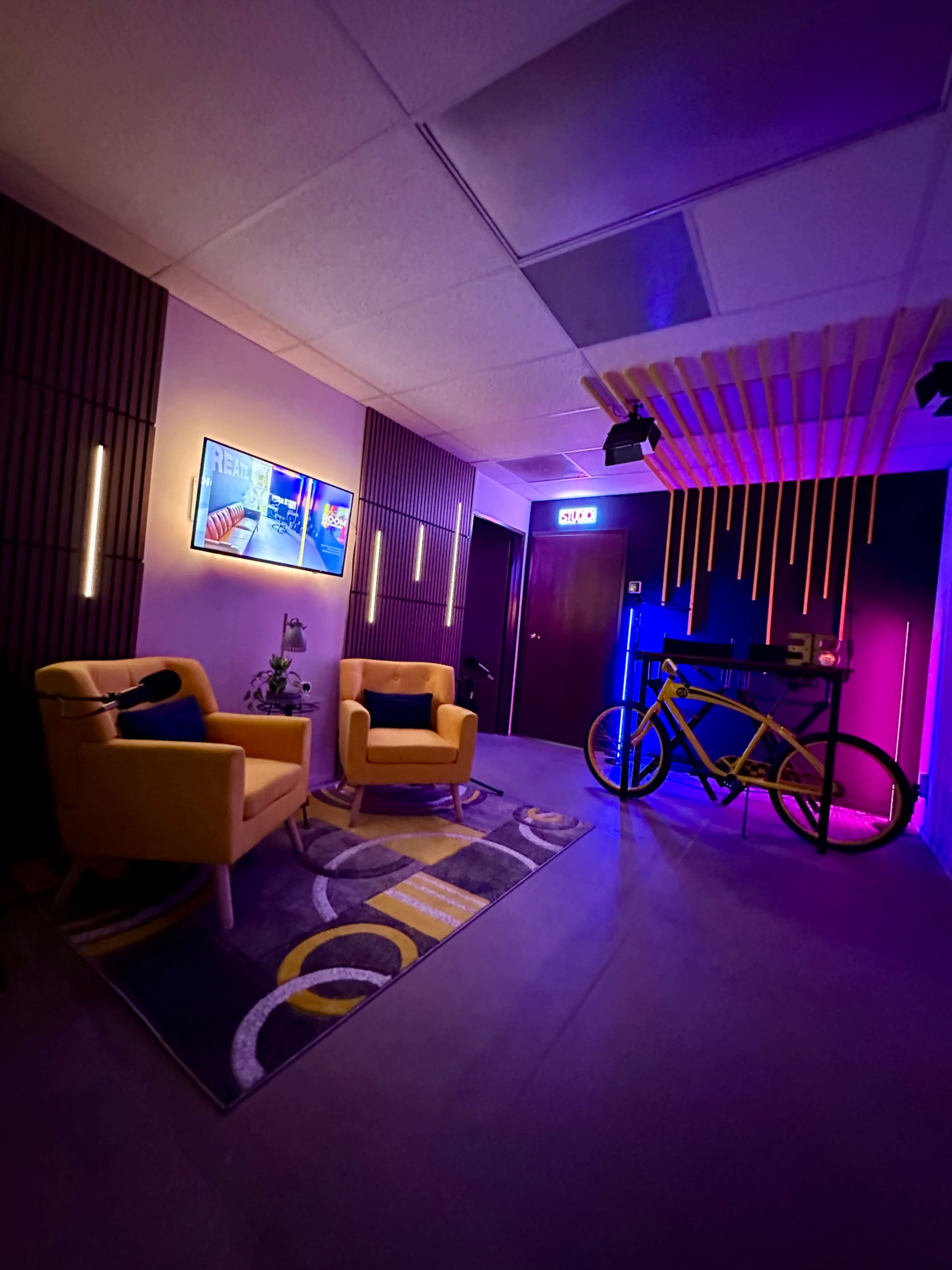 The image shows a modern lounge area featuring two yellow chairs, a bicycle, ambient lighting, and colorful wall decor.