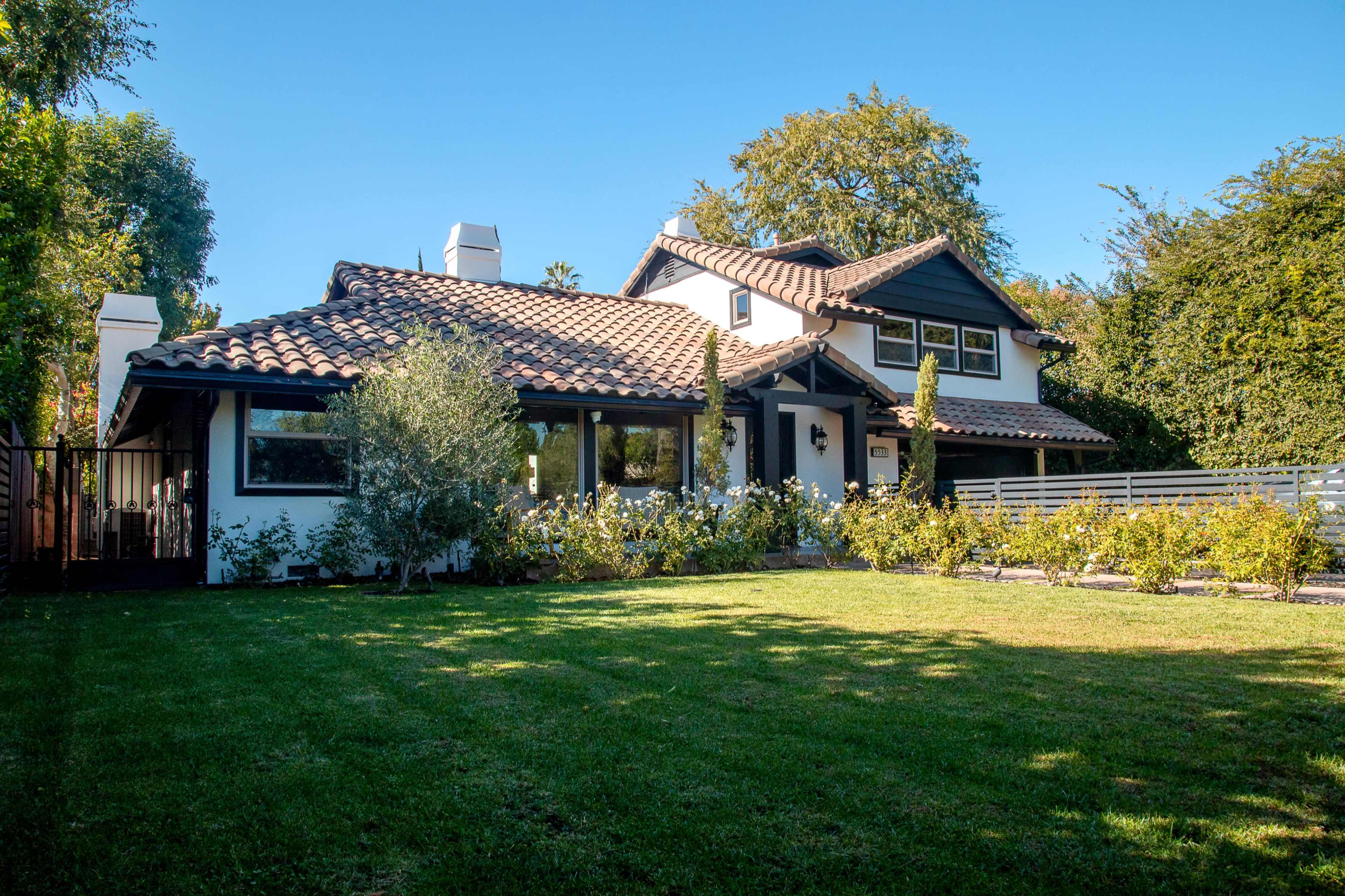 A two-story house with a tiled roof and white walls is surrounded by a green lawn and neatly trimmed bushes.