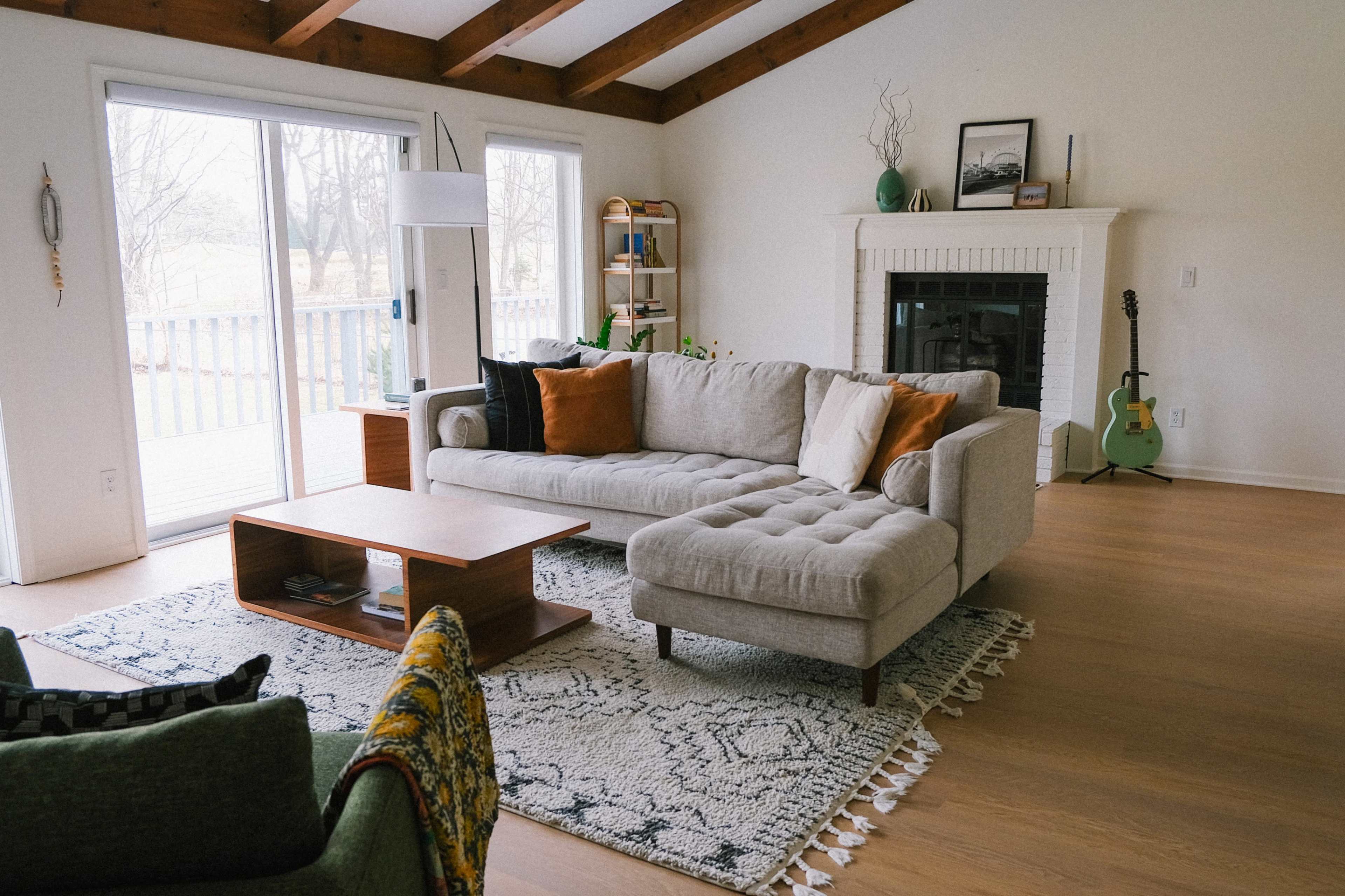 A spacious living room features a light gray sectional sofa with orange pillows, a wooden coffee table, and a guitar leaning against the wall next to a fireplace.