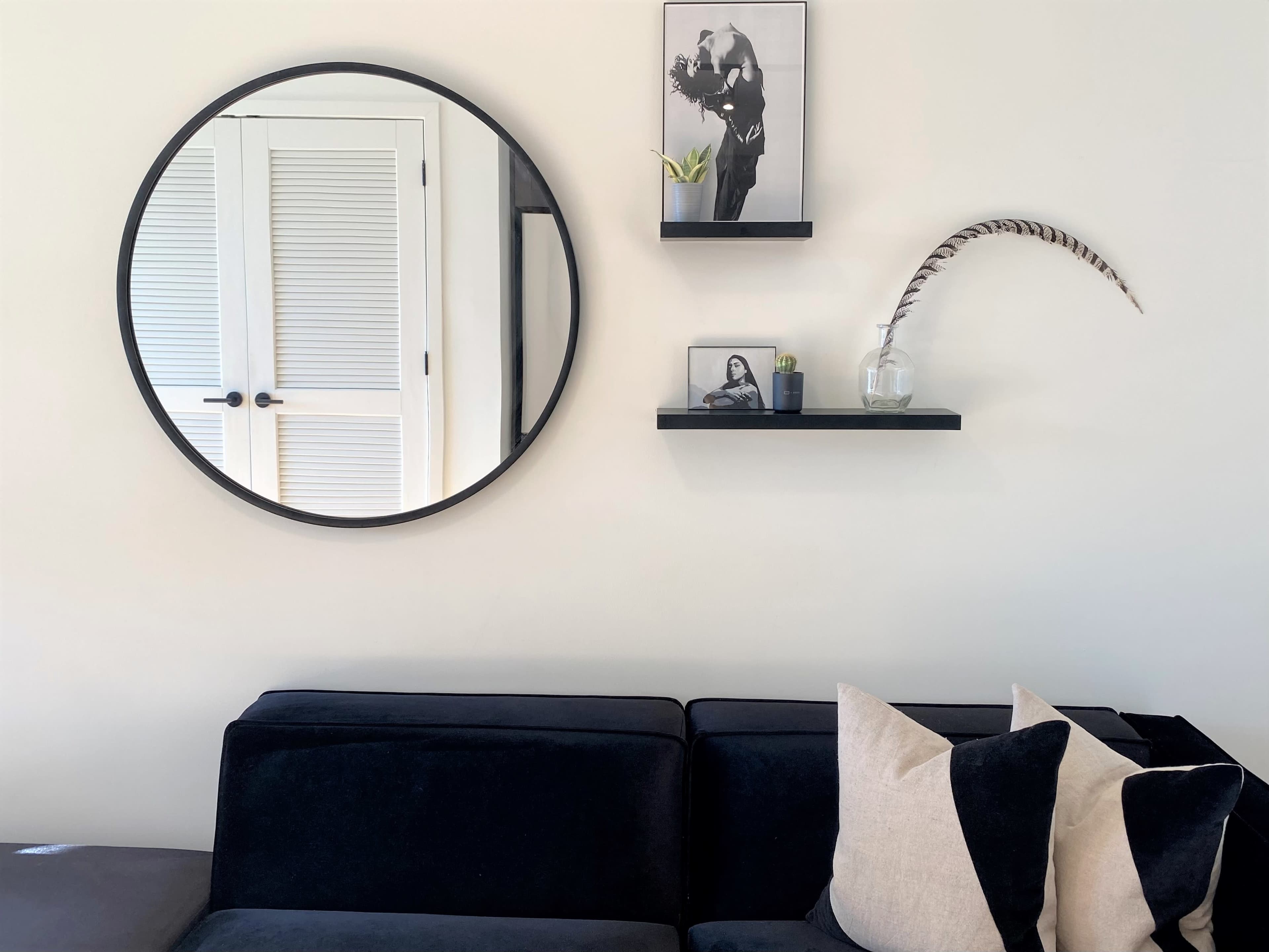 A round mirror hangs on a wall above a black sofa, accompanied by shelves displaying a photograph, a plant, and a decorative feather.
