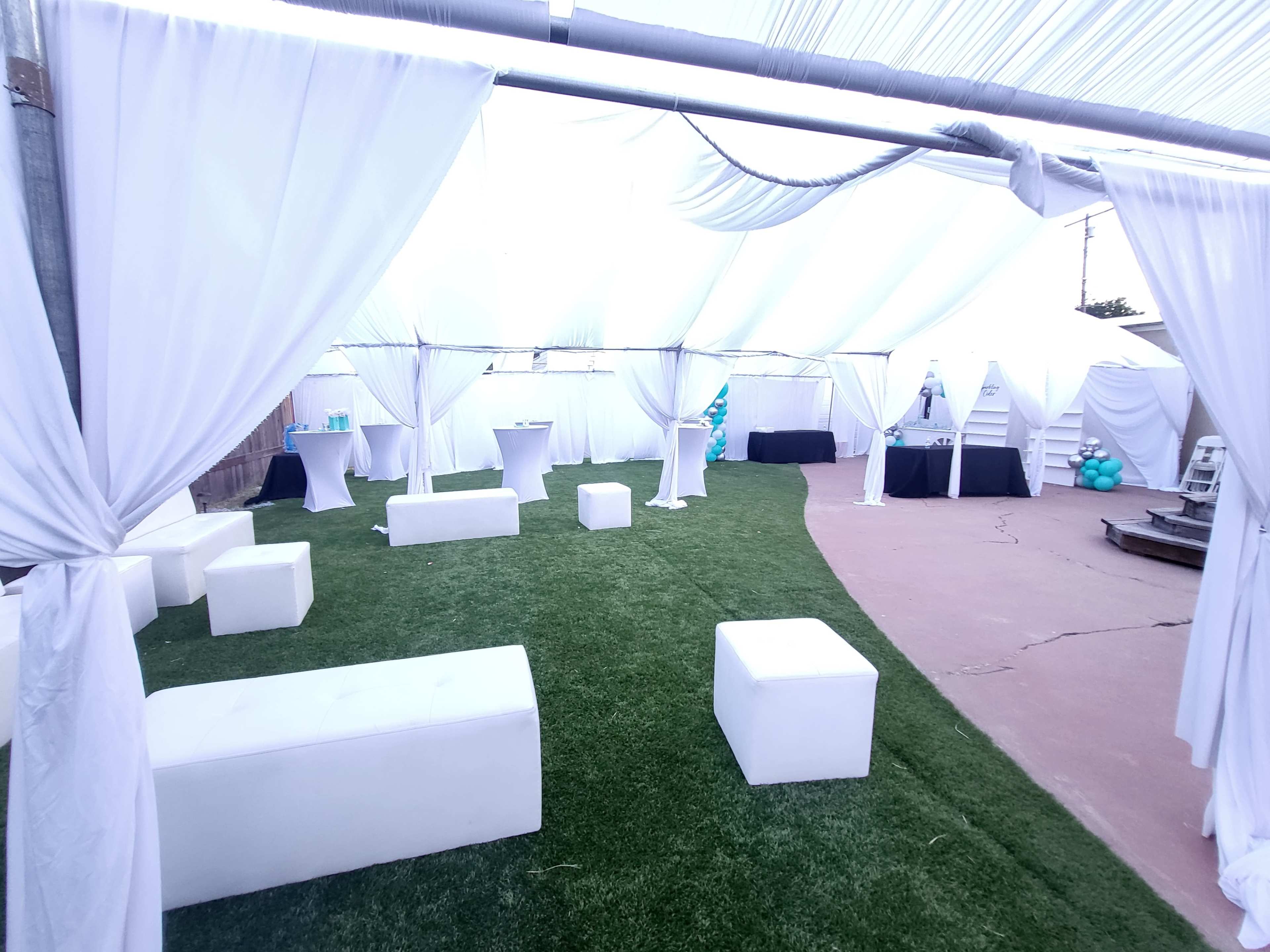 A tented outdoor event space featuring white draped fabric, green artificial grass, and various seating arrangements with cocktail tables and a black display table in the background.
