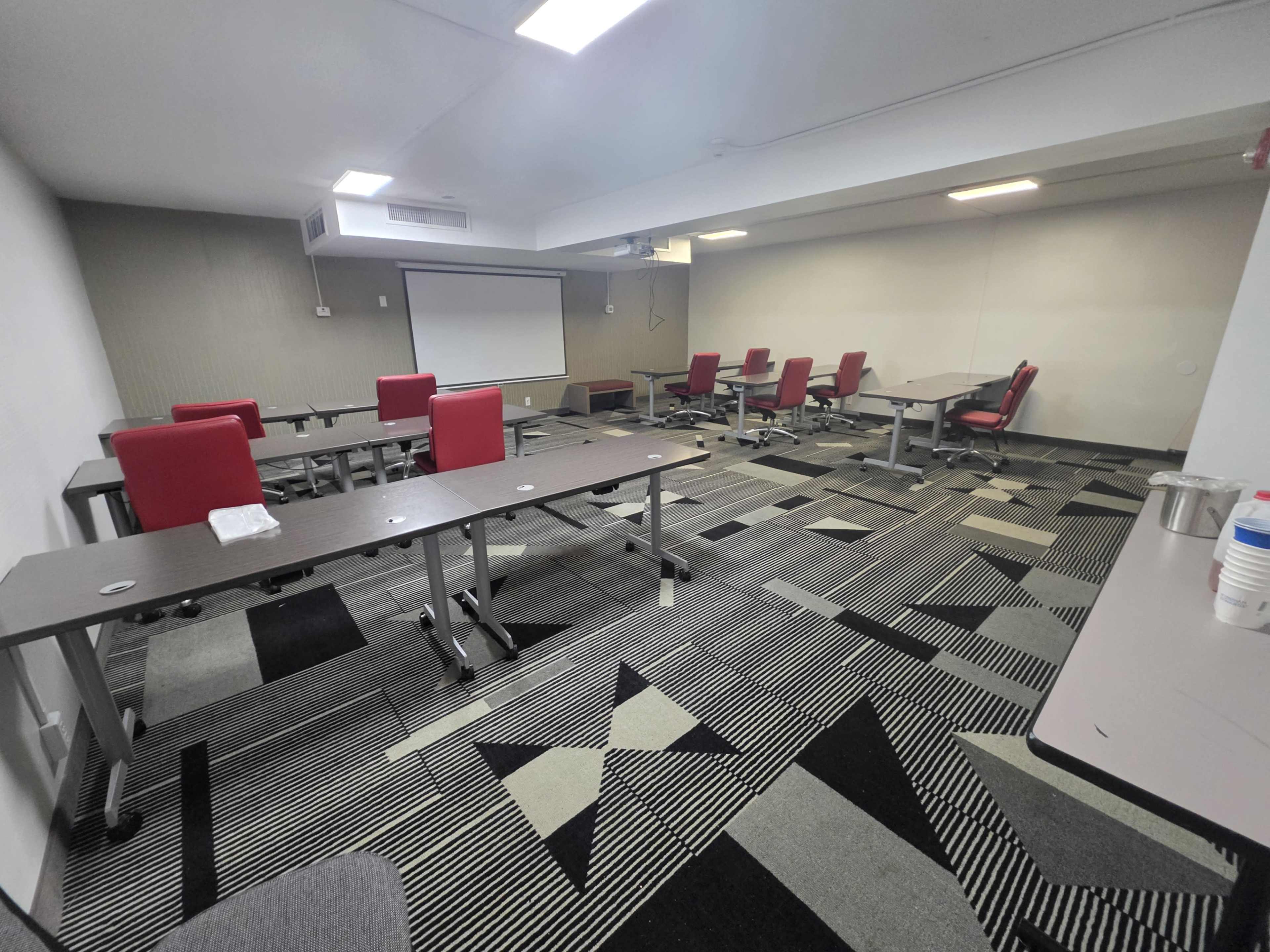 The image shows a conference room with several tables and chairs arranged in a sparse layout, along with a projector screen on the wall.