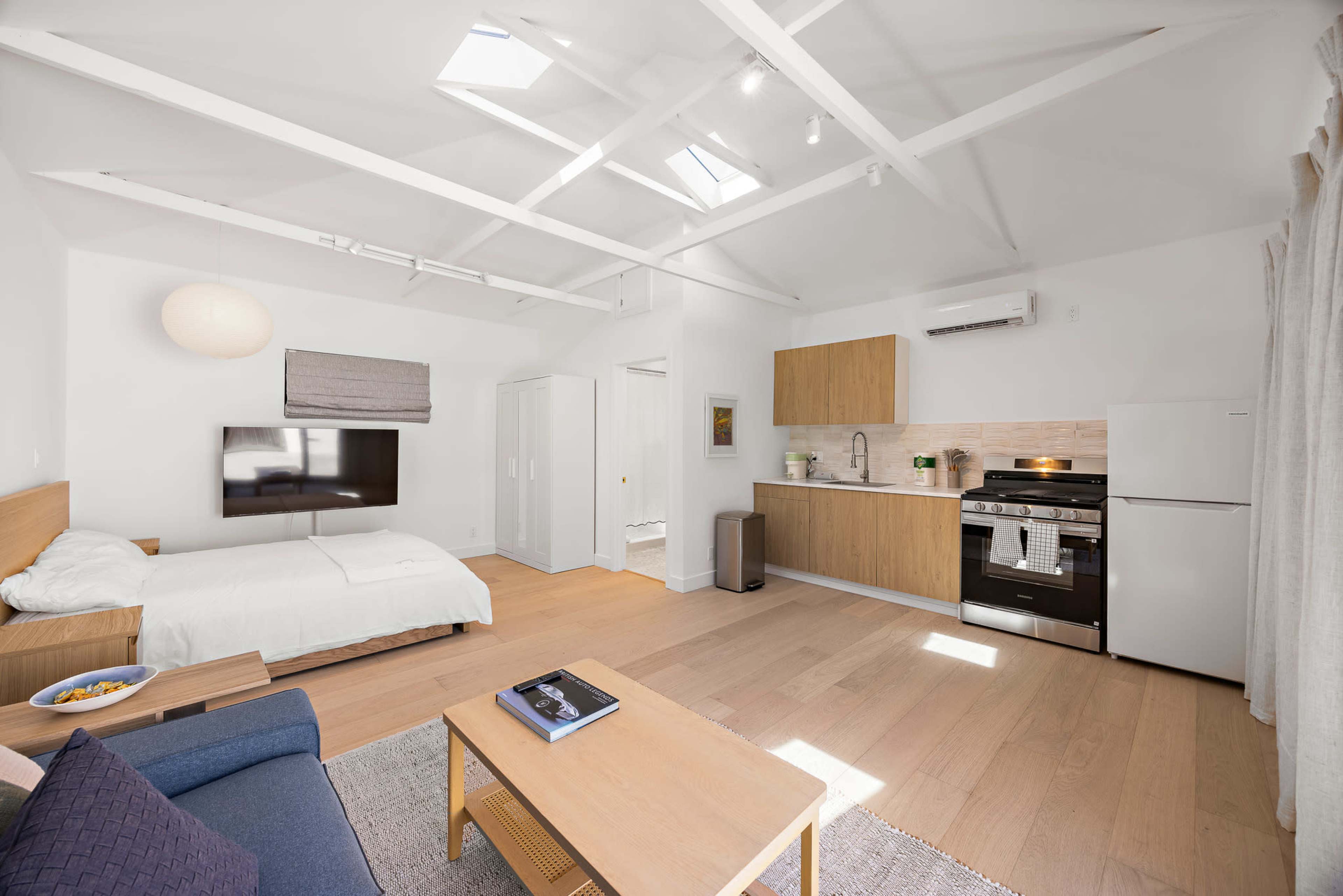 The image shows a modern studio apartment featuring a kitchen area, a living space with a couch, a bed, and large windows with skylights.