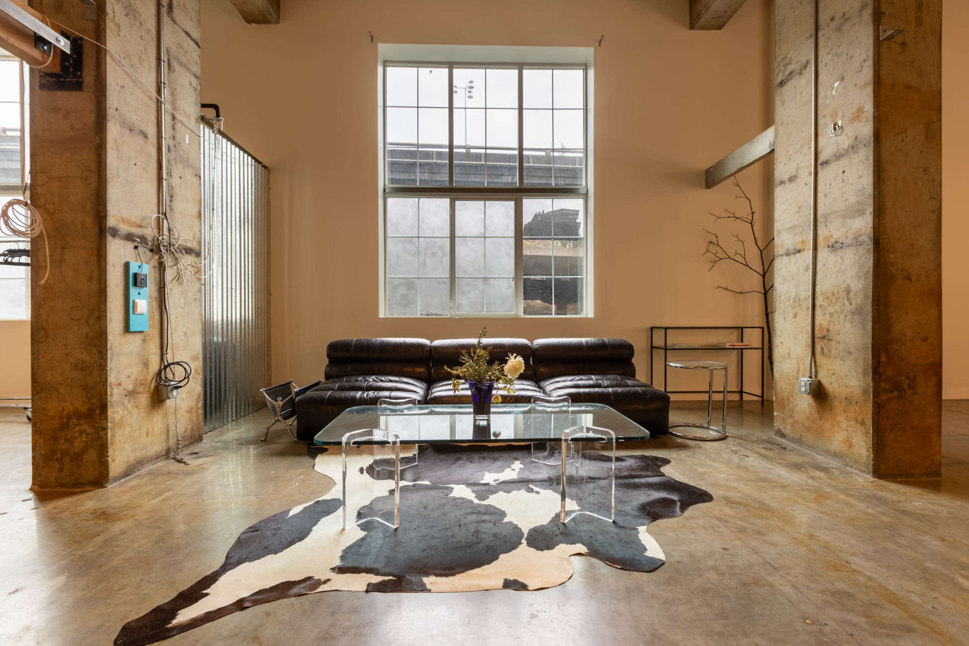 The image shows a modern, industrial-style living space featuring a black leather sofa, a glass coffee table on a cowhide rug, and large windows allowing natural light to enter.