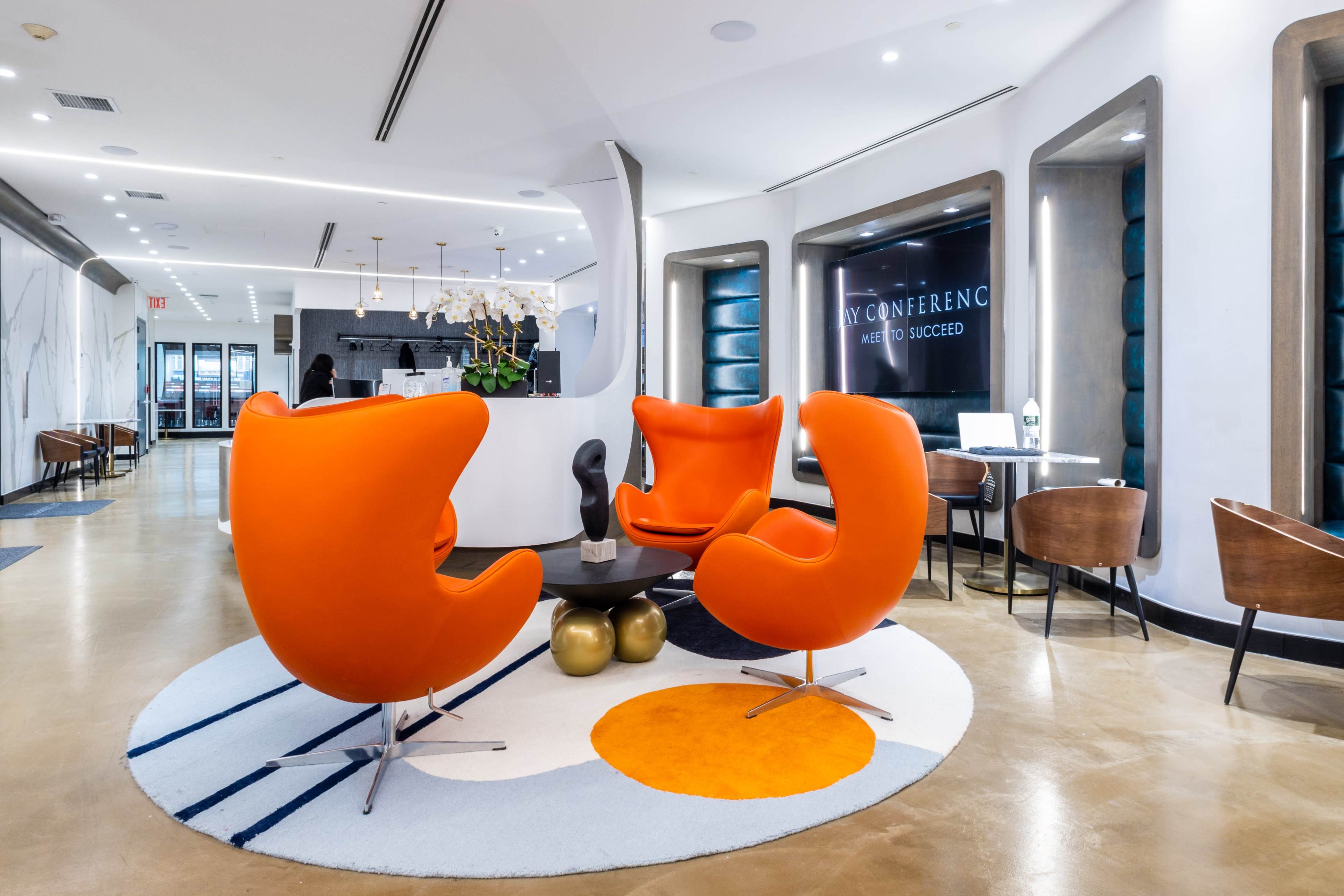 A contemporary lounge area with two orange chairs, a circular rug featuring geometric patterns, and a reception desk in a well-lit space.