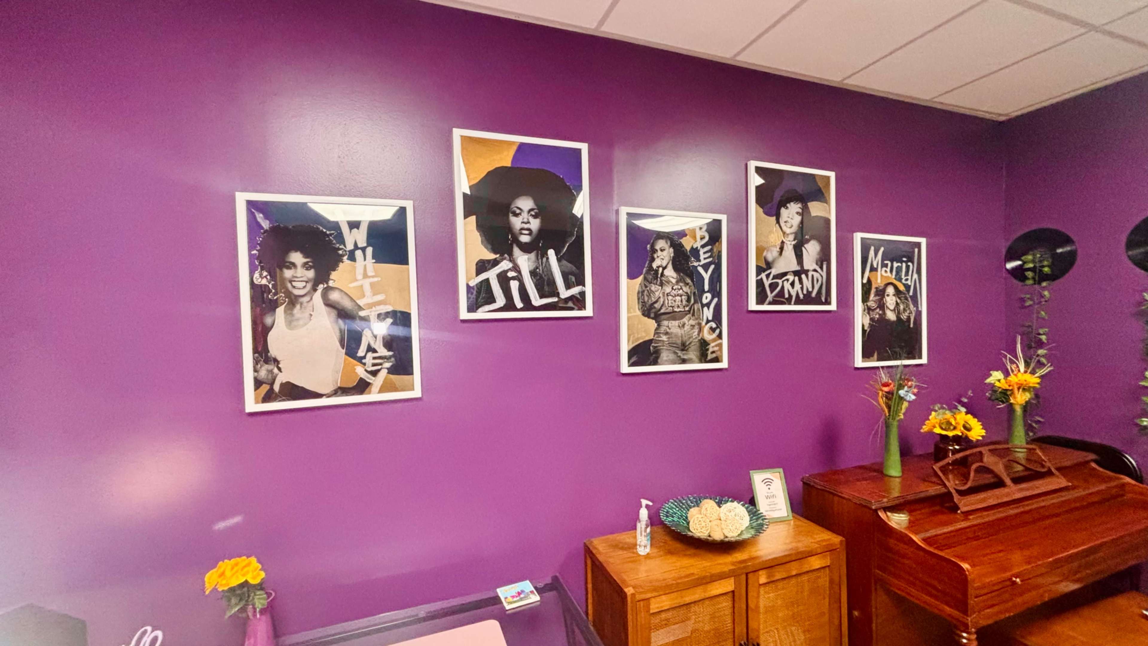The image shows a purple wall adorned with five framed portraits of women, accompanied by flowers on a wooden table in front.