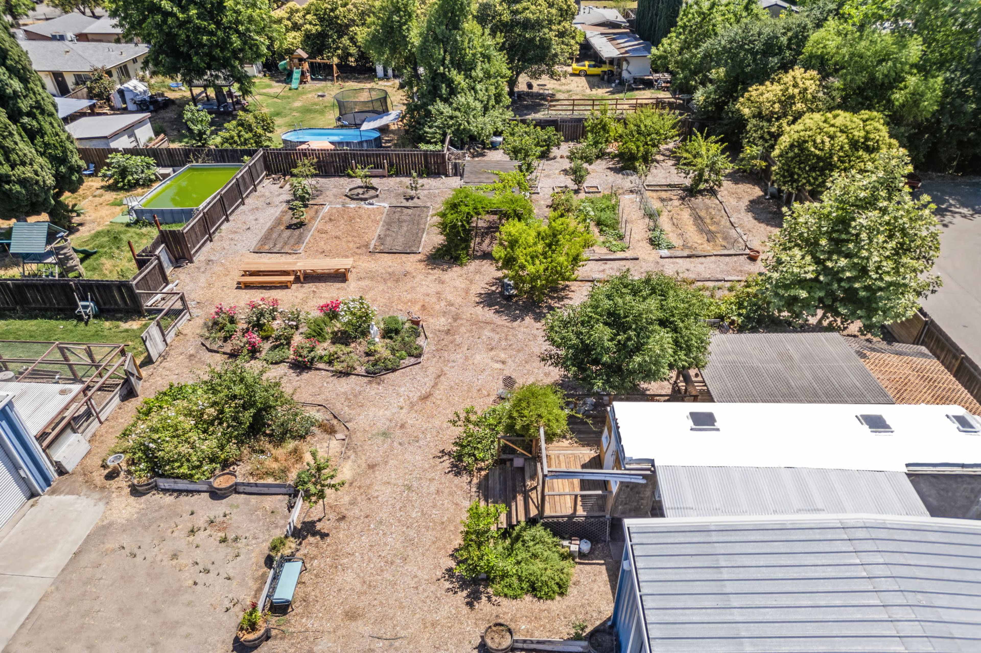 An aerial view of a spacious backyard garden featuring various planting beds, trees, and seating areas, surrounded by a wooden fence.