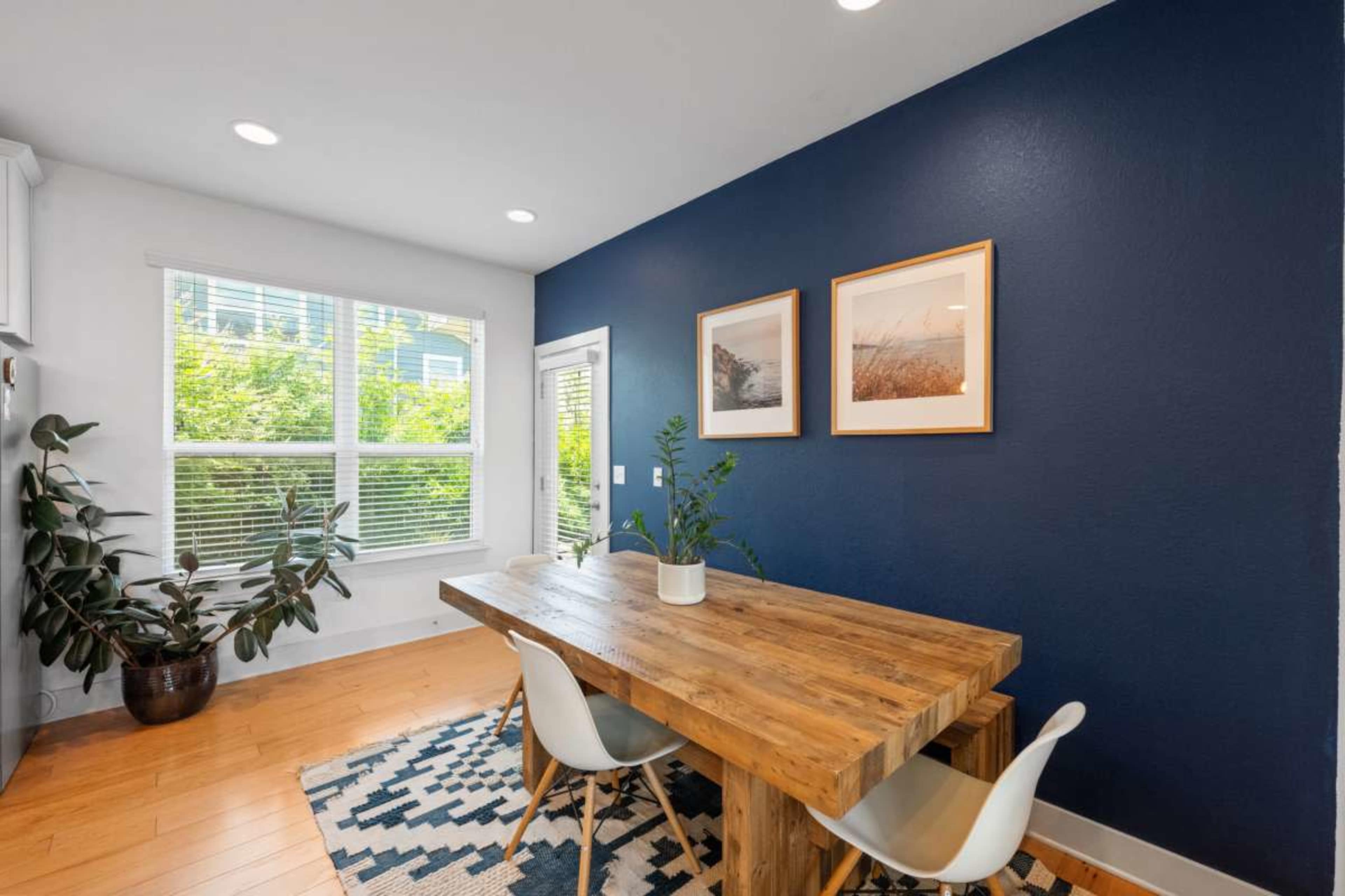 A wooden dining table with white chairs sits on a patterned rug in a brightly lit room featuring a blue accent wall and large windows.