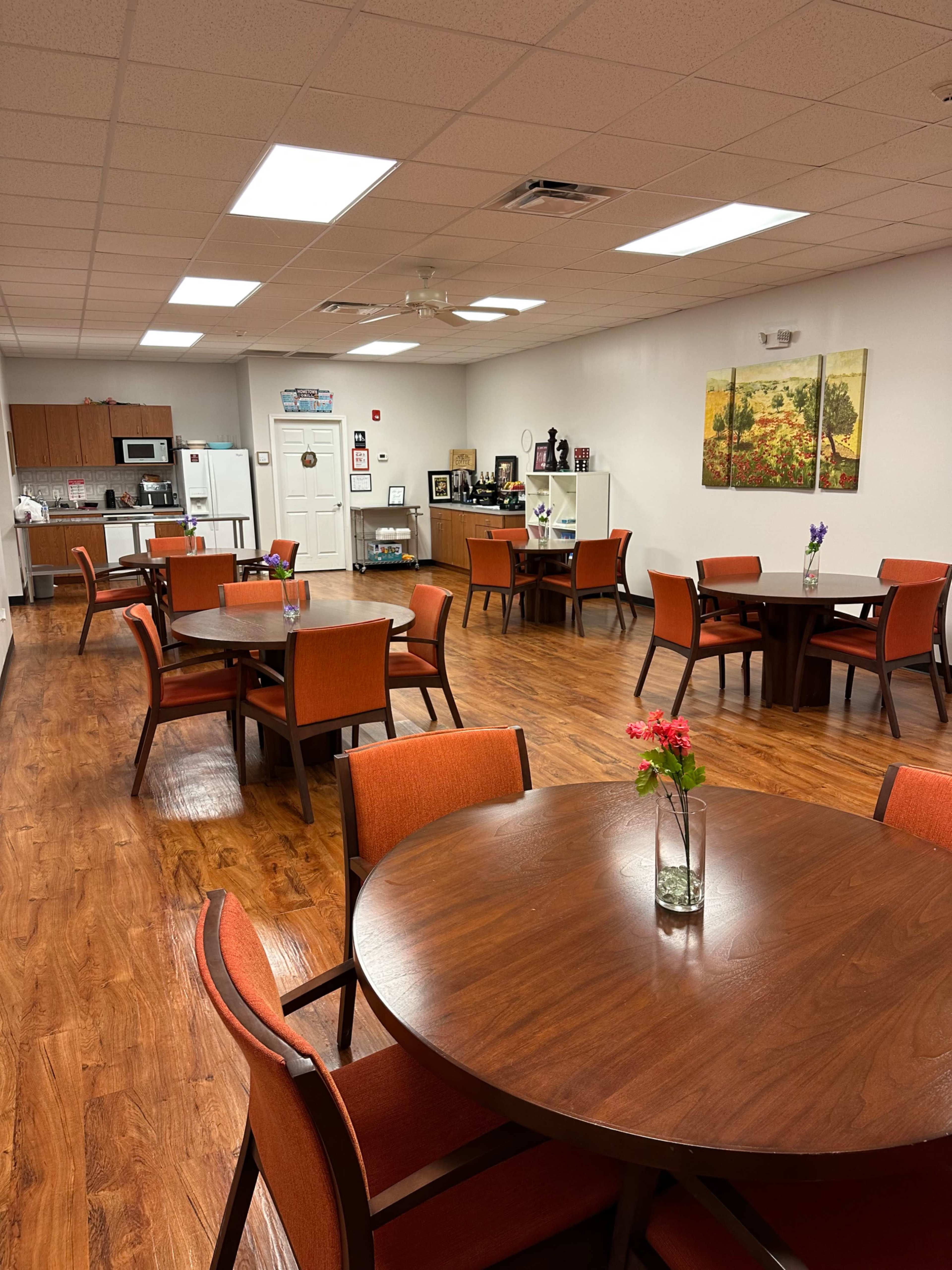 A dining area with wooden tables and orange chairs, equipped with a kitchenette in the background.