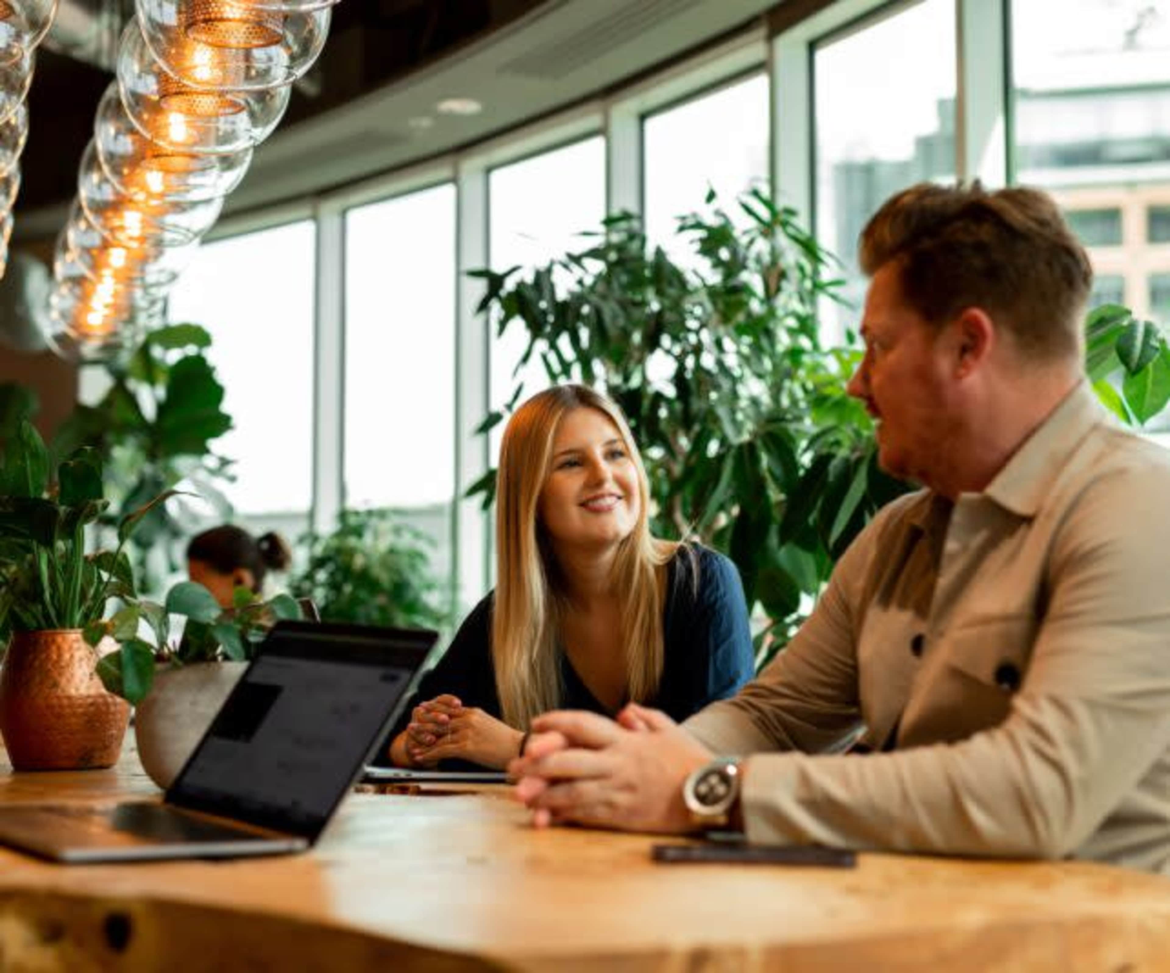 Two individuals are seated at a wooden table in a modern workspace, surrounded by large windows and greenery, engaging in conversation.