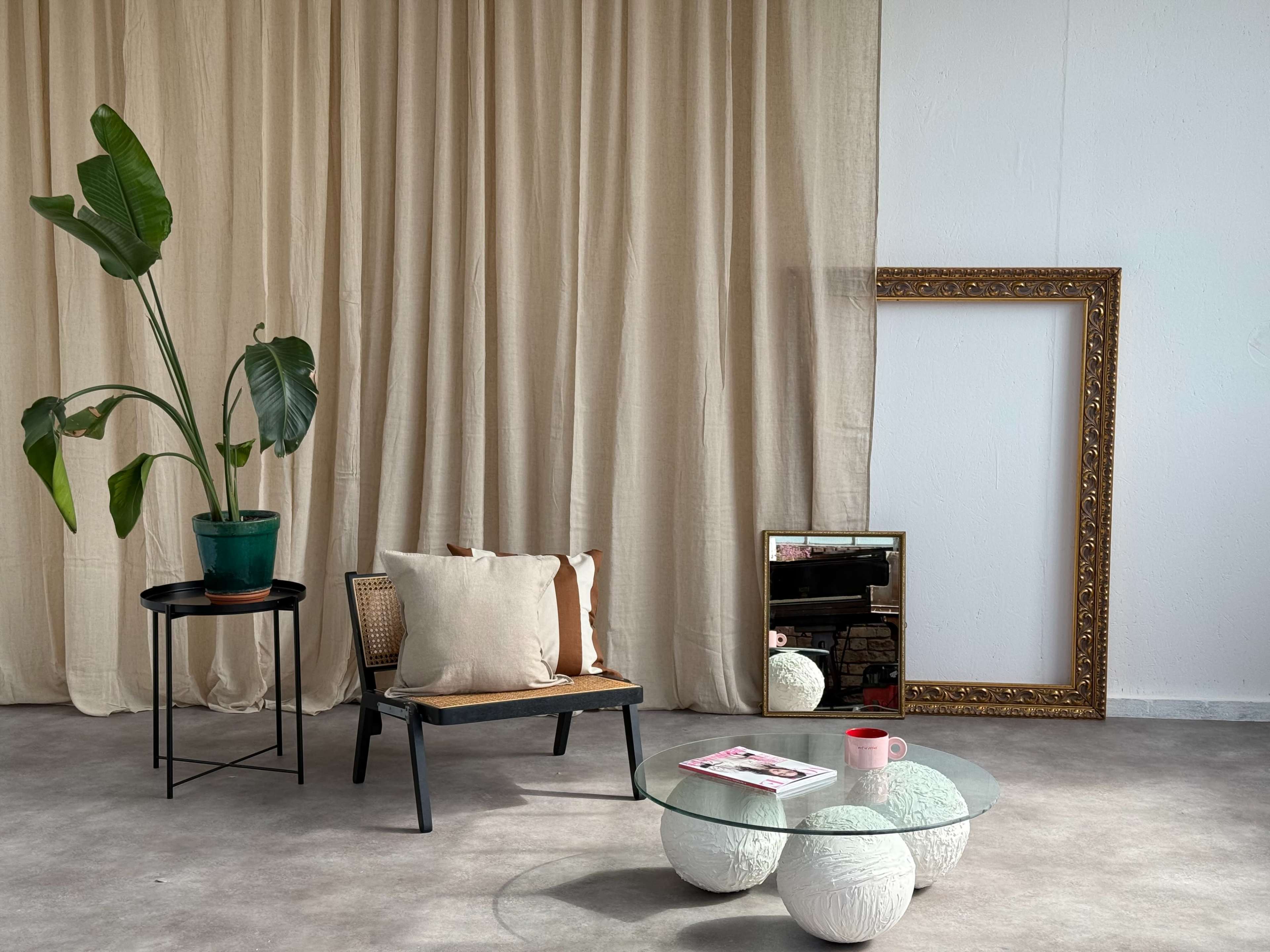 A minimalist interior scene features a black chair with cushions, a round glass coffee table, a potted plant, a standing ornate mirror, and a beige curtain backdrop.
