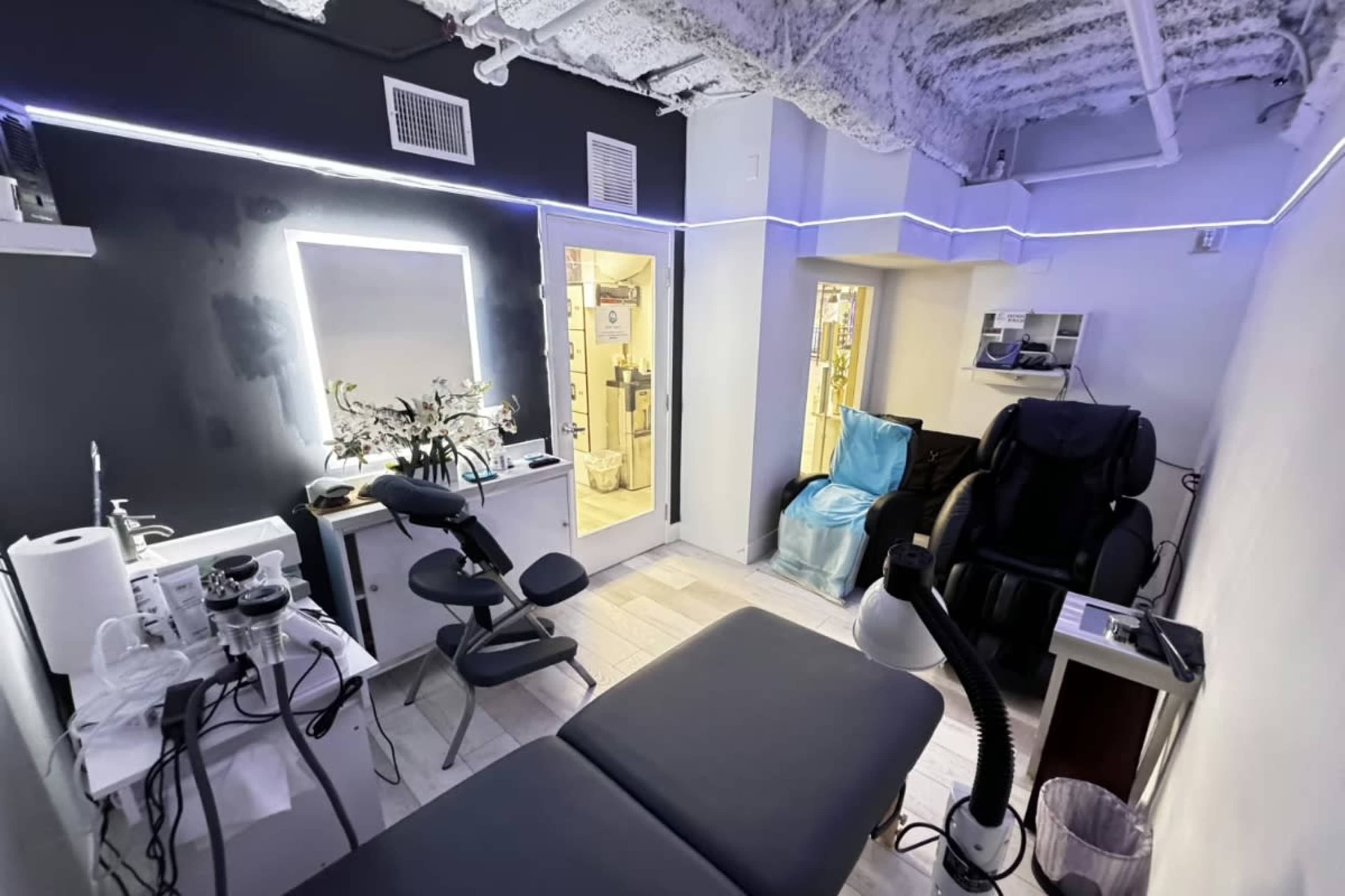 The image shows a modern treatment room with a massage table, various equipment, and a lighted mirror, along with chairs and a door leading to another area.