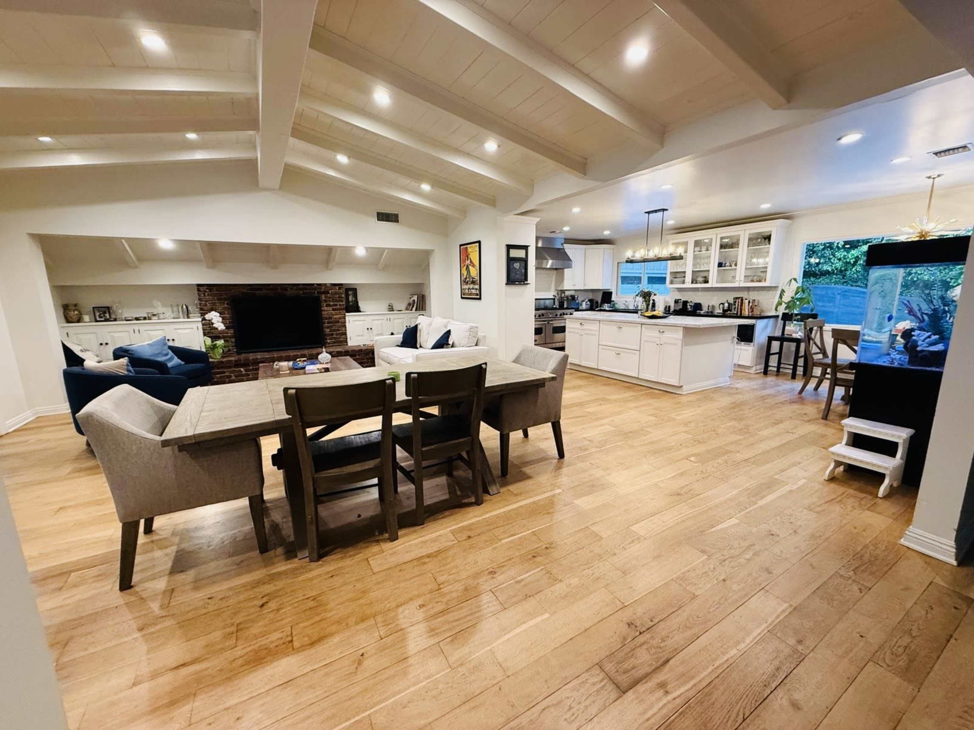 The image shows a spacious, inviting living area with a dining table, comfortable seating, and an open kitchen, featuring wooden floors and exposed beam ceilings.