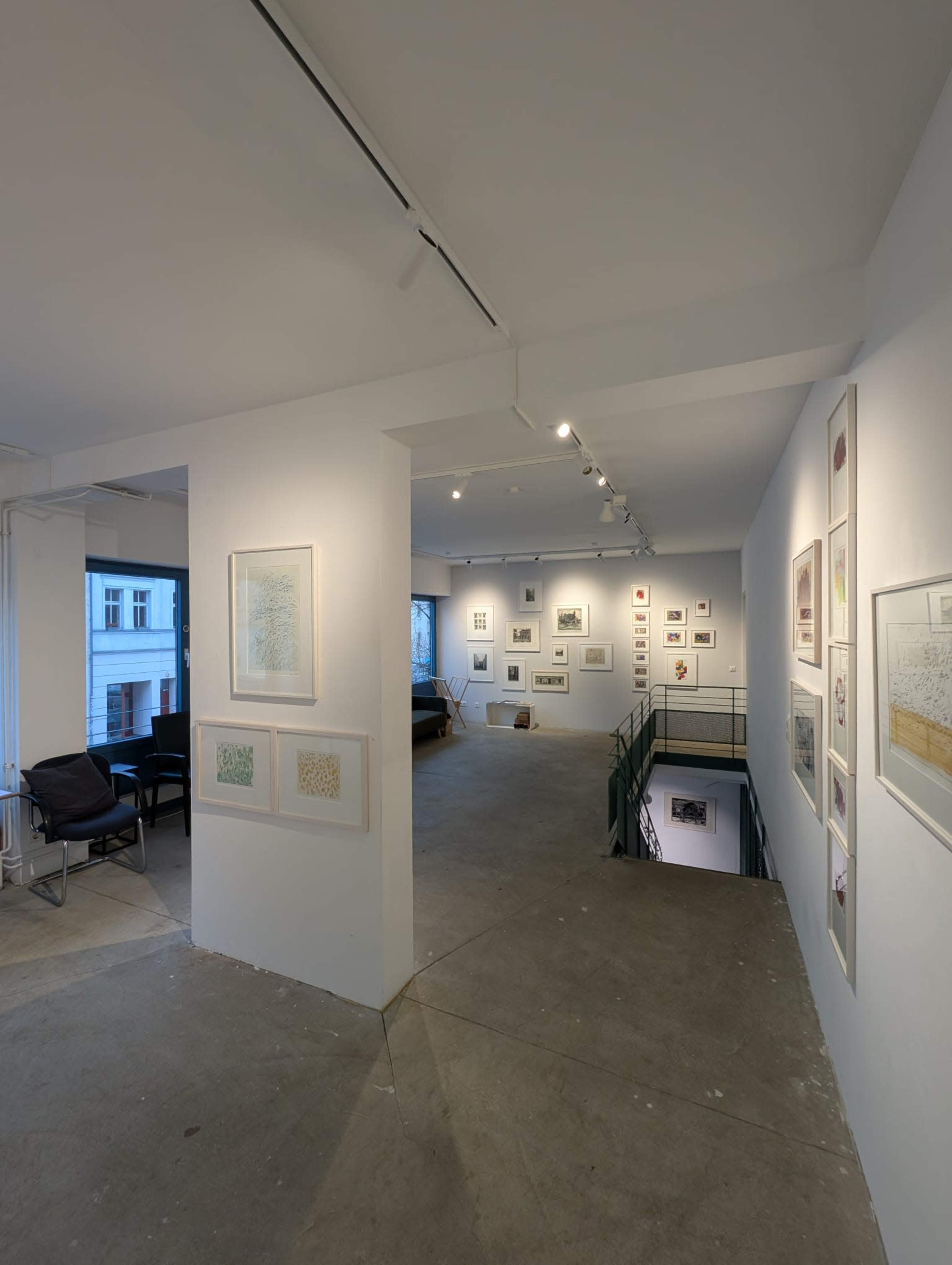 The image shows an interior view of an art gallery with white walls displaying various artworks and seating in the foreground.
