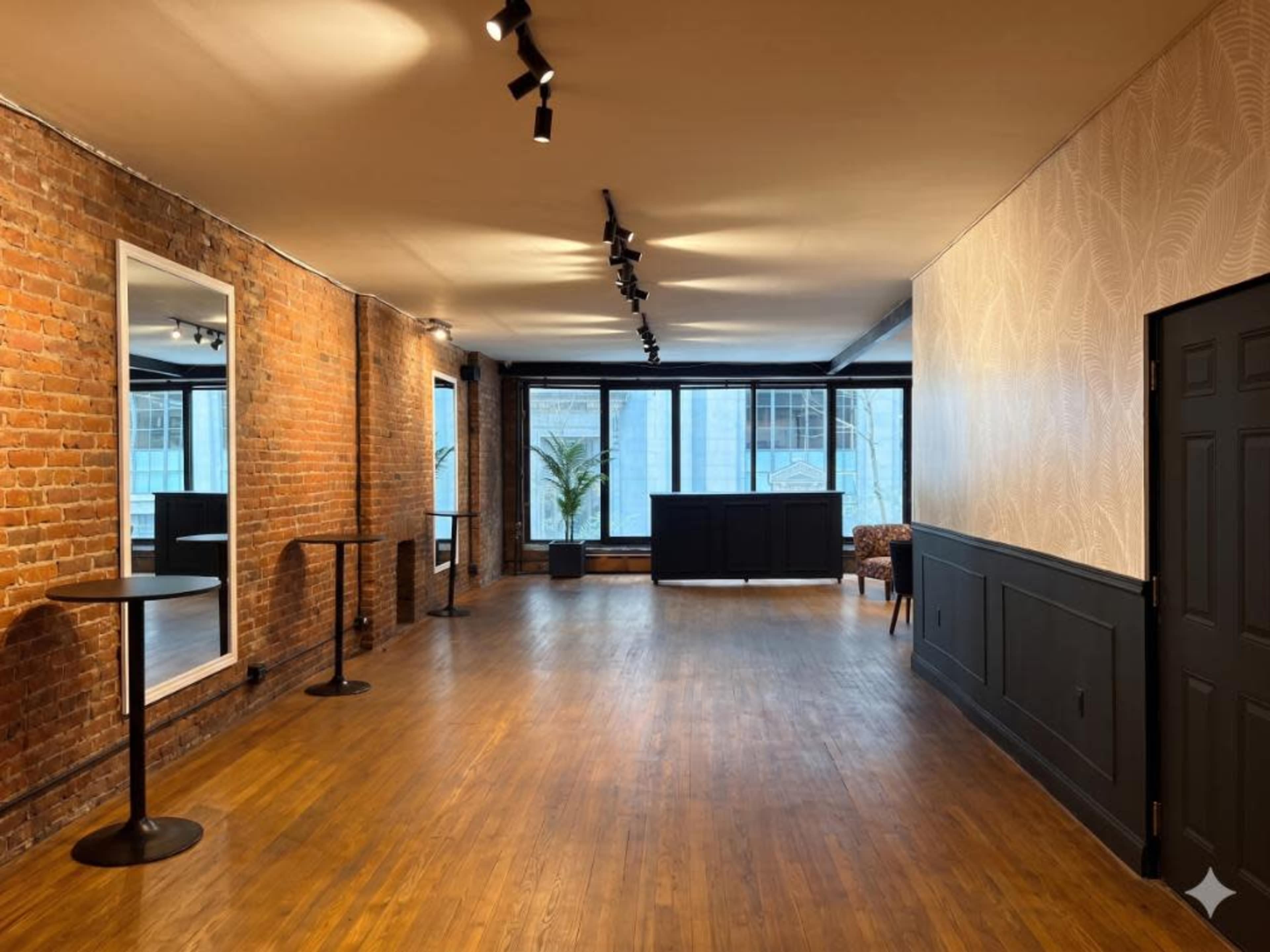 The image shows a spacious interior with exposed brick walls, wooden flooring, and a reception area featuring a dark counter and minimal furniture.
