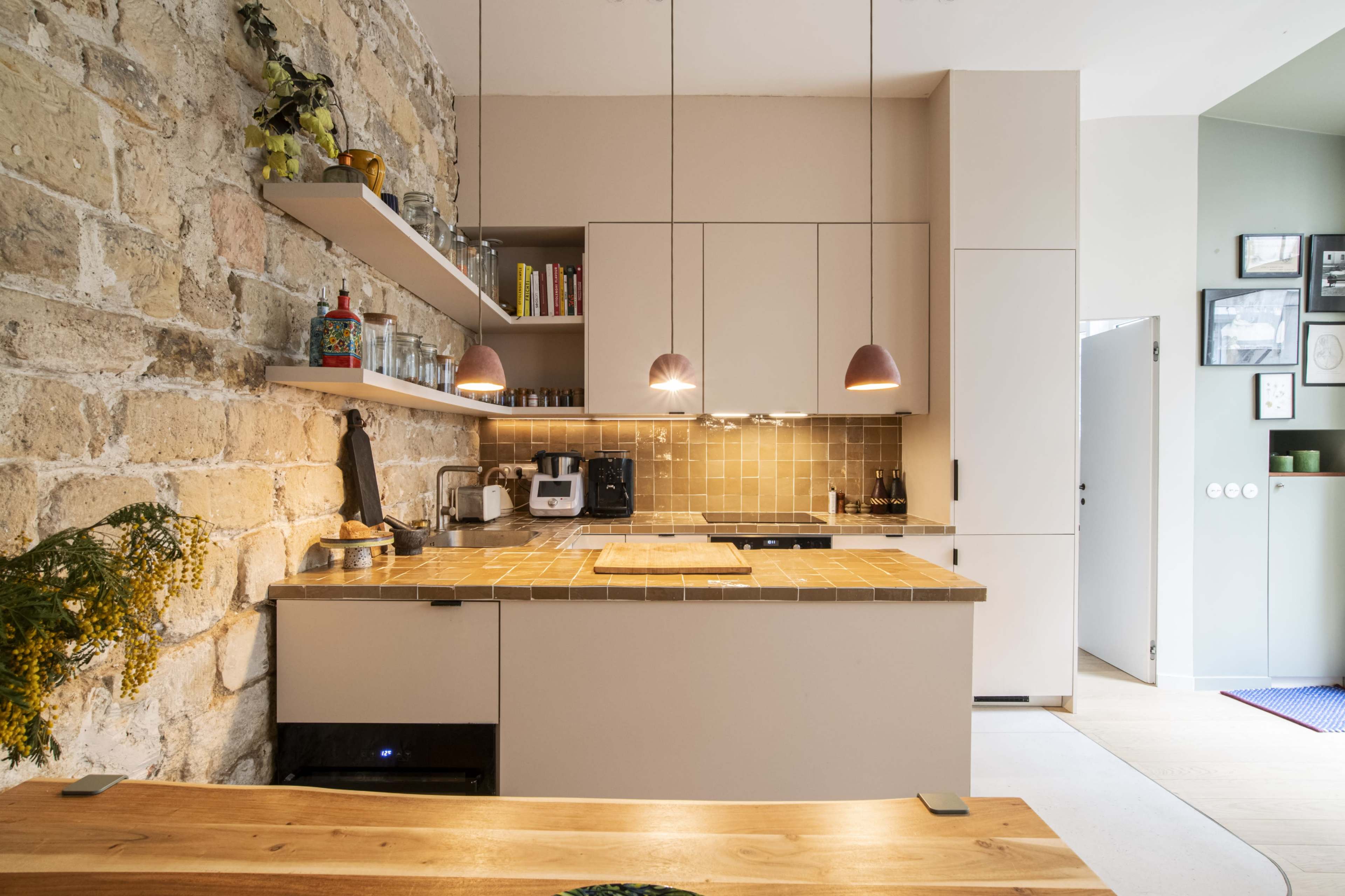 The image shows a modern kitchen with a stone wall, wooden countertops, and minimalist cabinetry, featuring pendant lights above the counter.