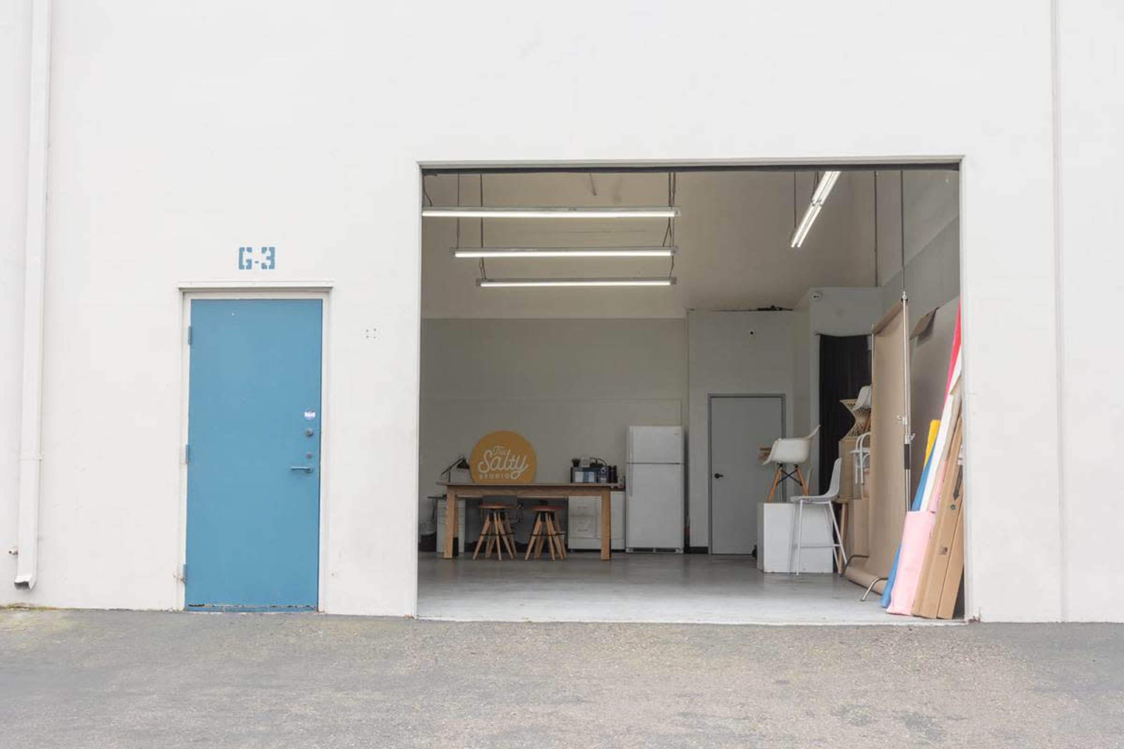The image shows the exterior of a warehouse space with an open garage door revealing a minimalist interior that includes a table, chairs, and kitchen appliances.