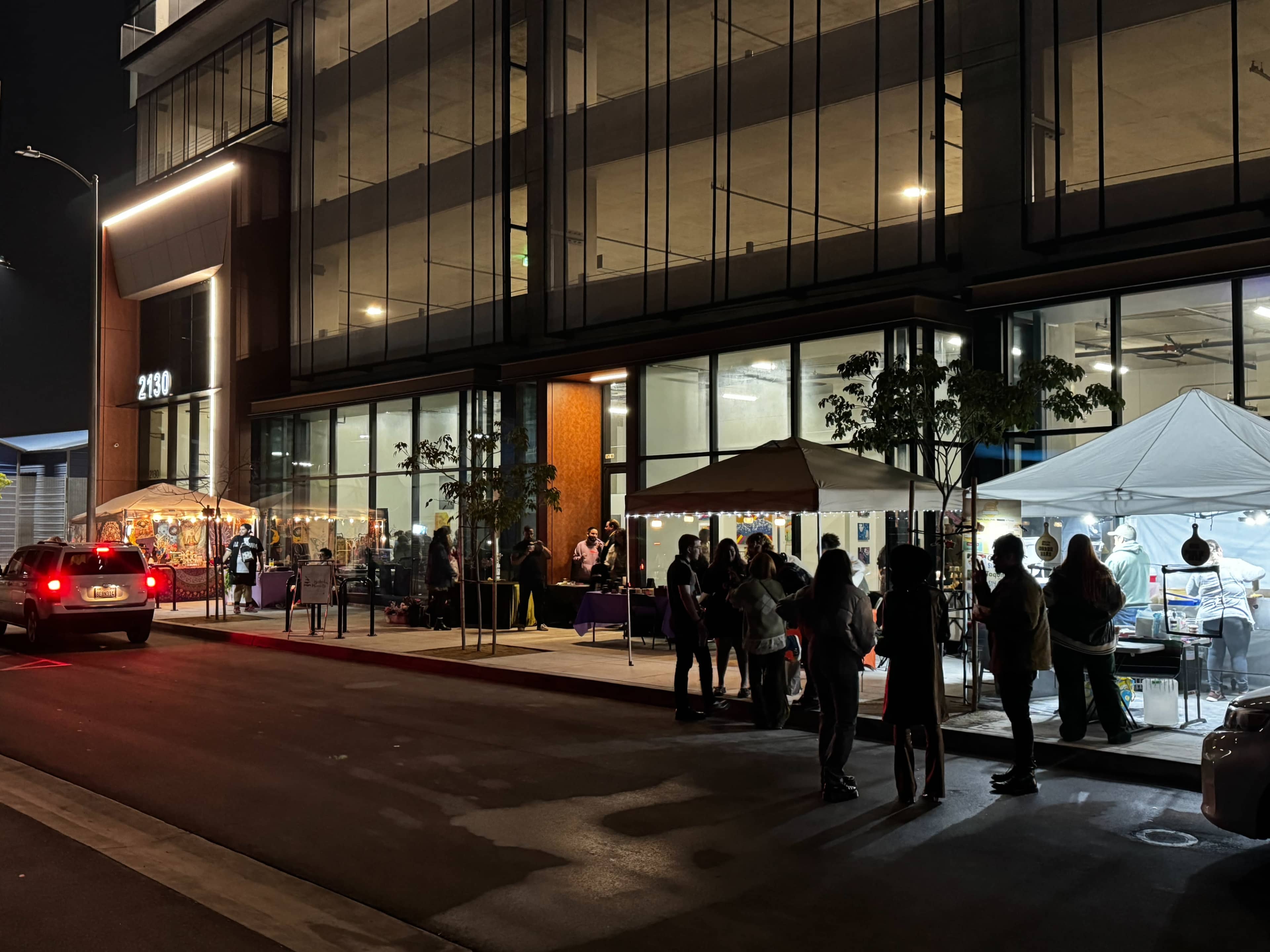 A group of people stands in line outside a building with large windows, while food vendors set up their stalls nearby under tents.