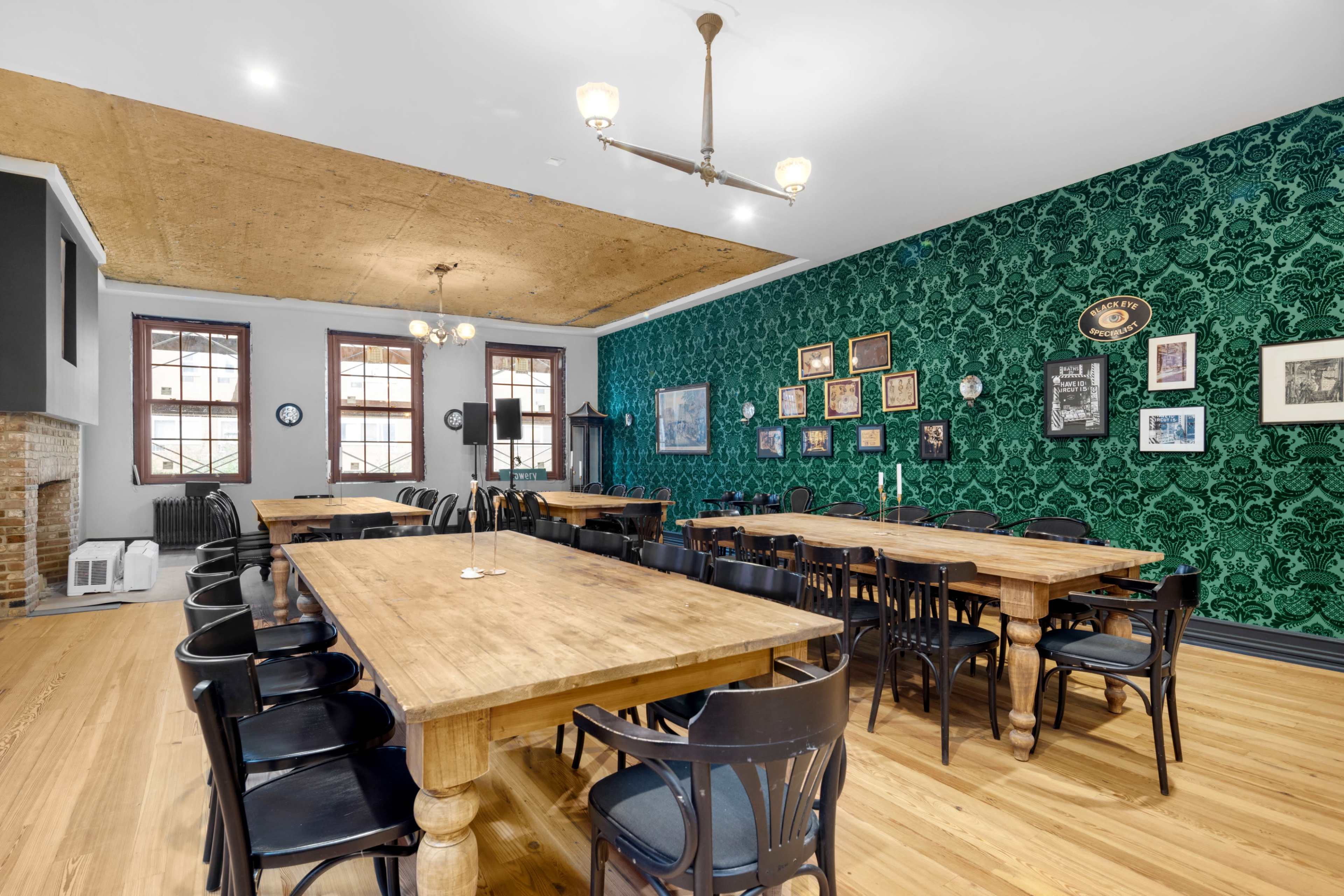 A spacious dining room with wooden tables and black chairs, featuring green patterned wallpaper and various framed pictures on the walls.