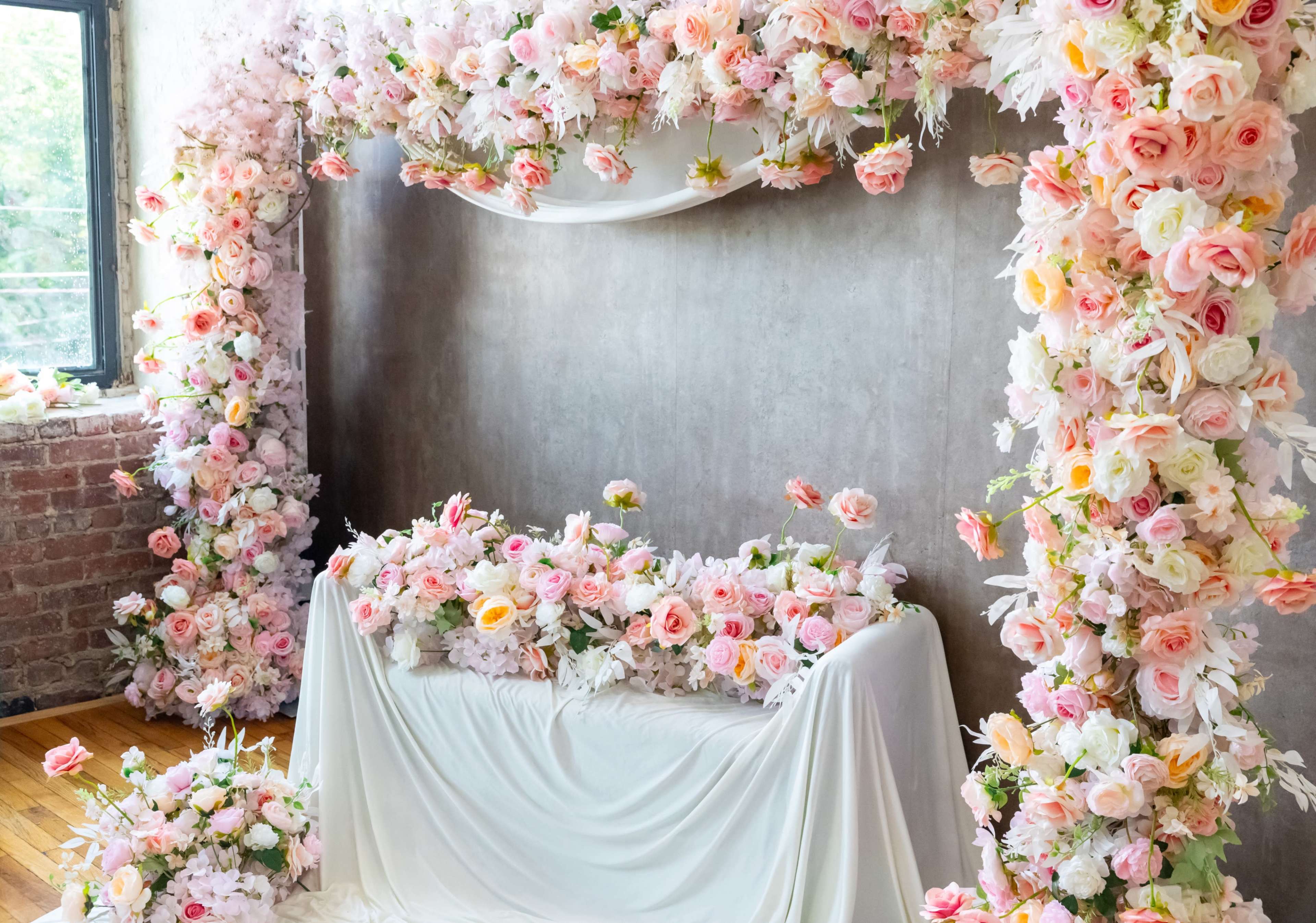 A floral installation featuring an arch and a table adorned with various roses and other flowers in shades of pink and white.