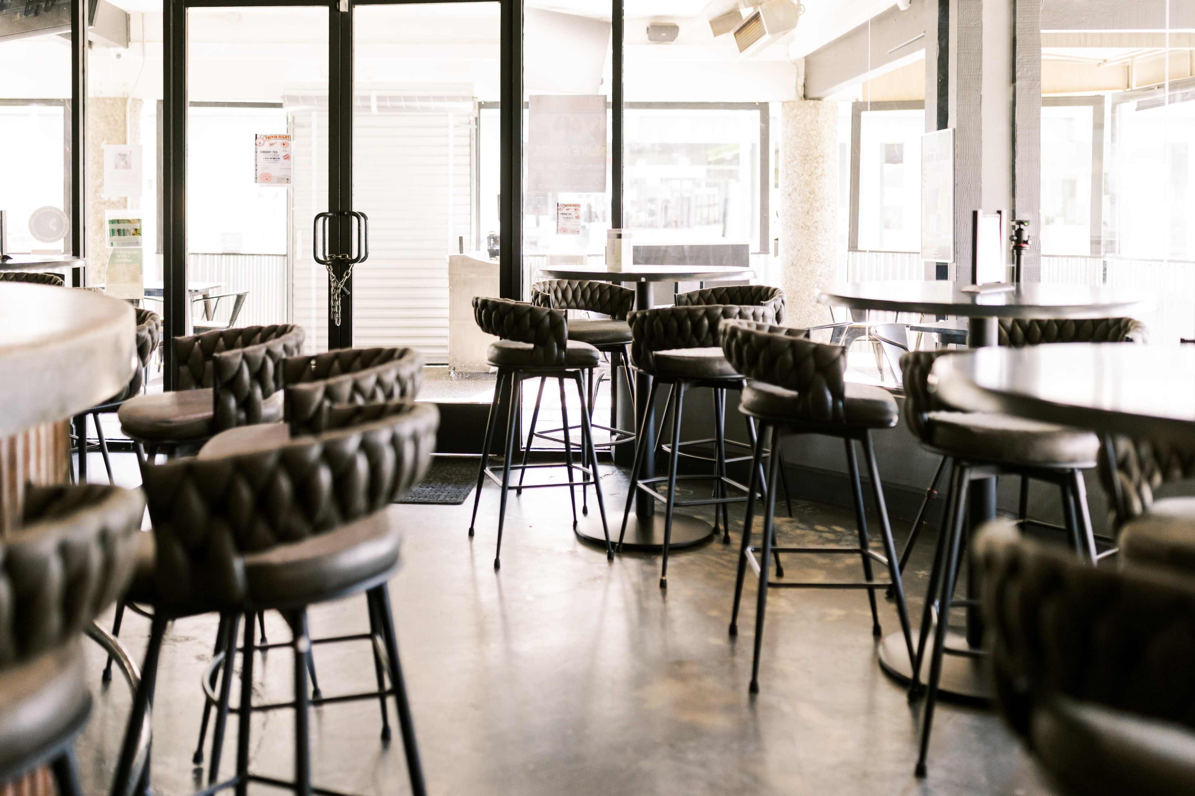 The image shows a modern bar area with high seating, empty tables, and large windows allowing natural light to enter.