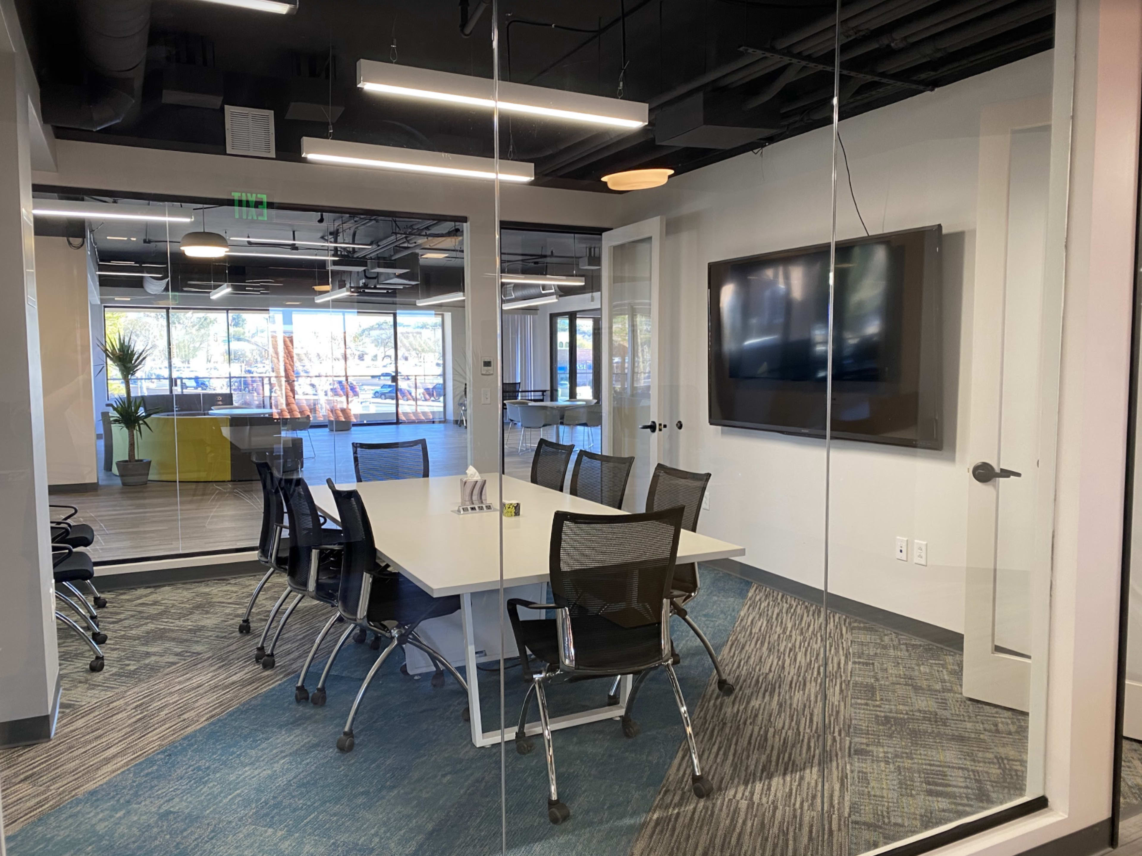 A glass-walled conference room equipped with a large table, chairs, and a flat-screen TV.