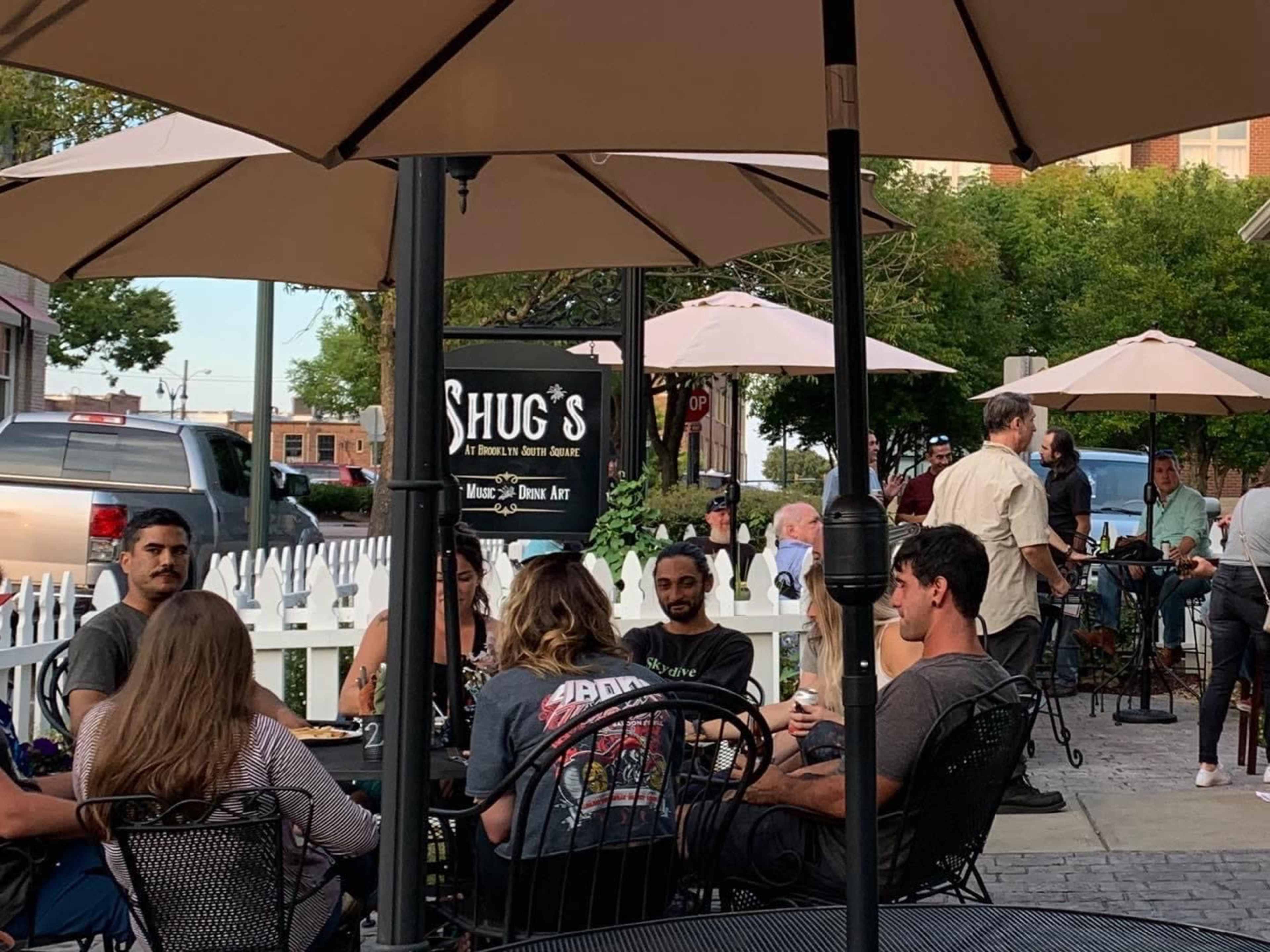 A group of people sits at outdoor tables under umbrellas at a restaurant called Shug's, with a variety of vehicles parked nearby.