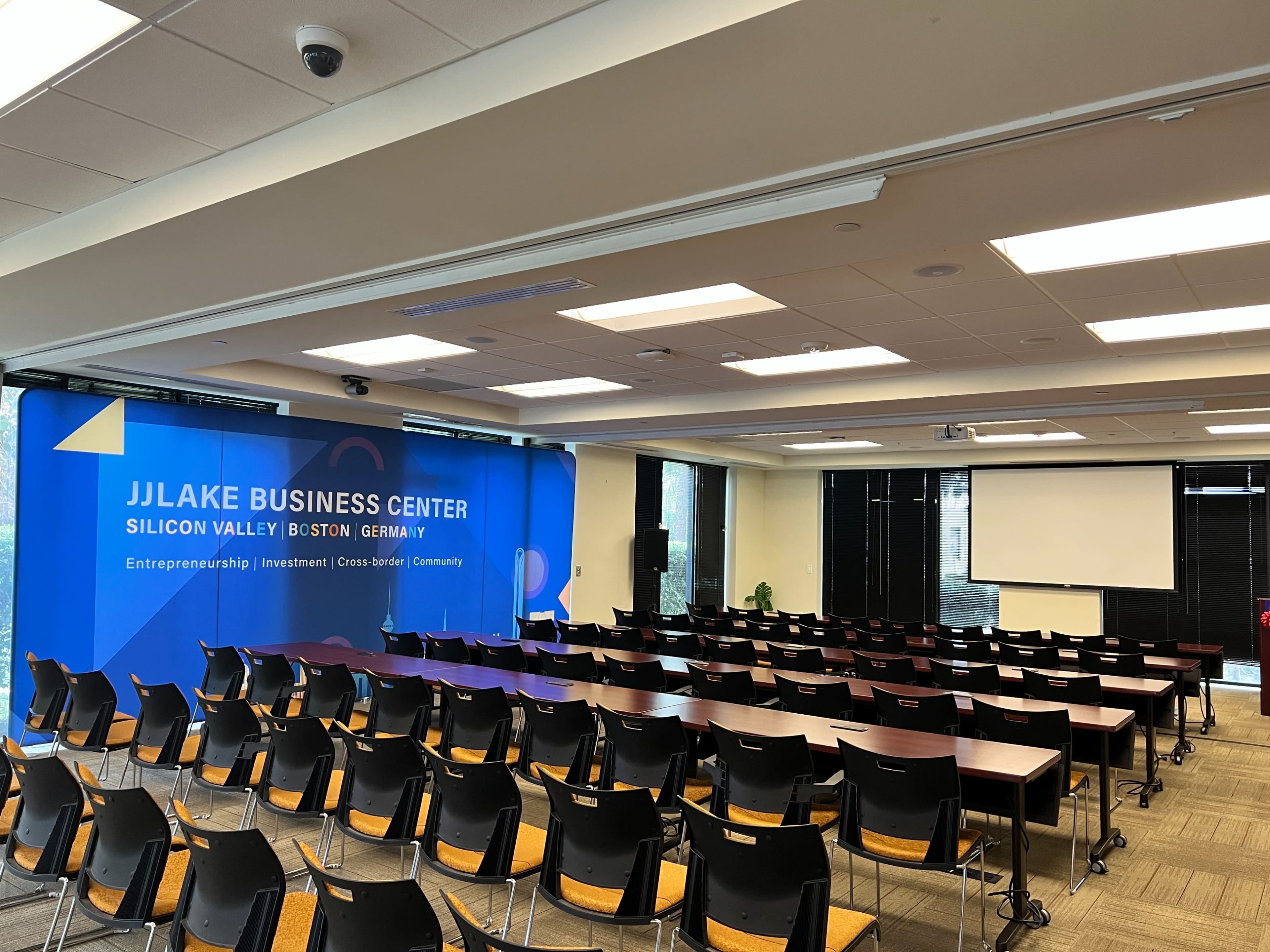 A seminar room at the JJLake Business Center, featuring rows of tables and chairs facing a presentation area with a large backdrop.