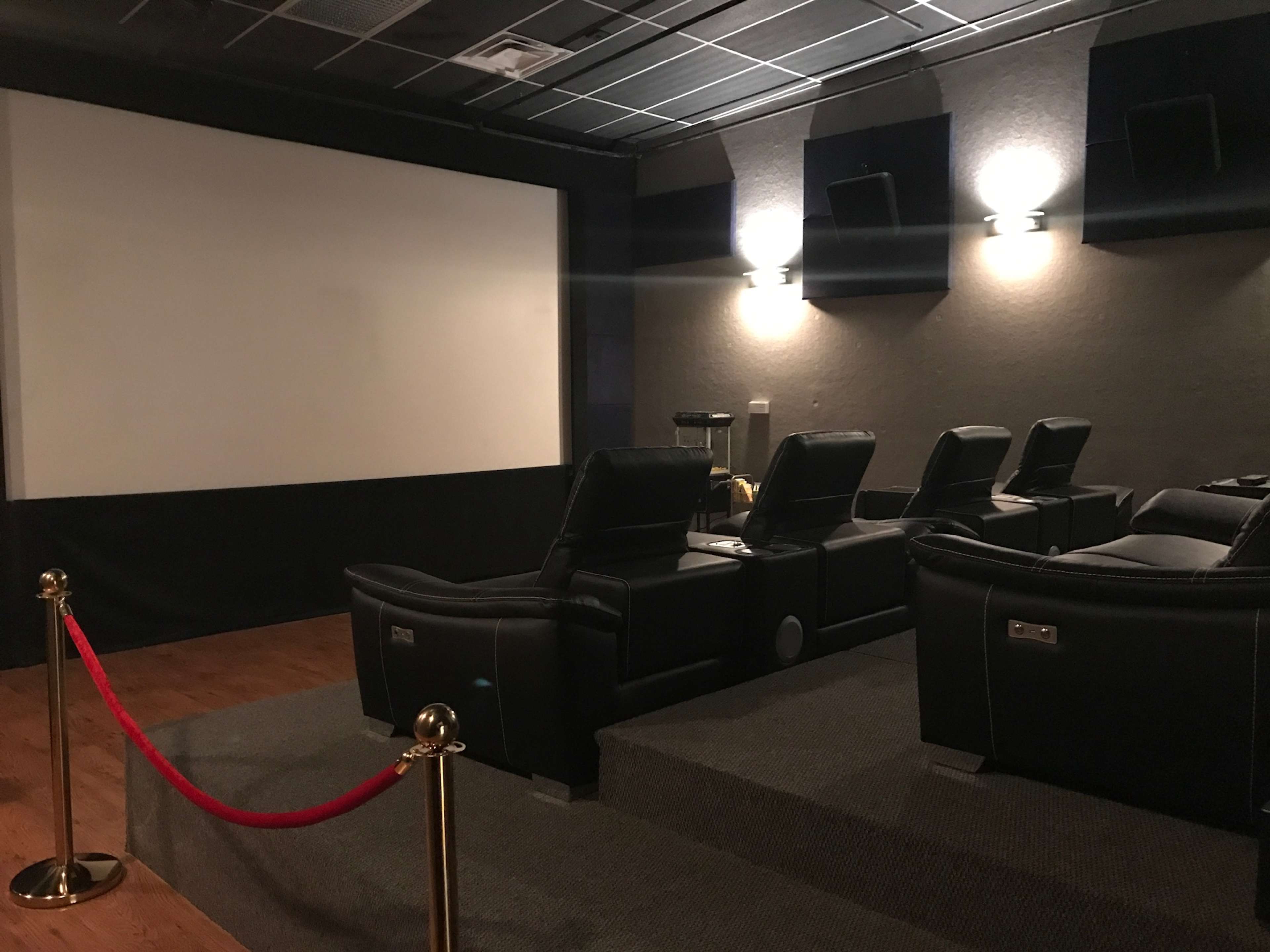 A small home theater with reclining black seats, a large screen at the front, and red ropes separating the seating area from the entrance.