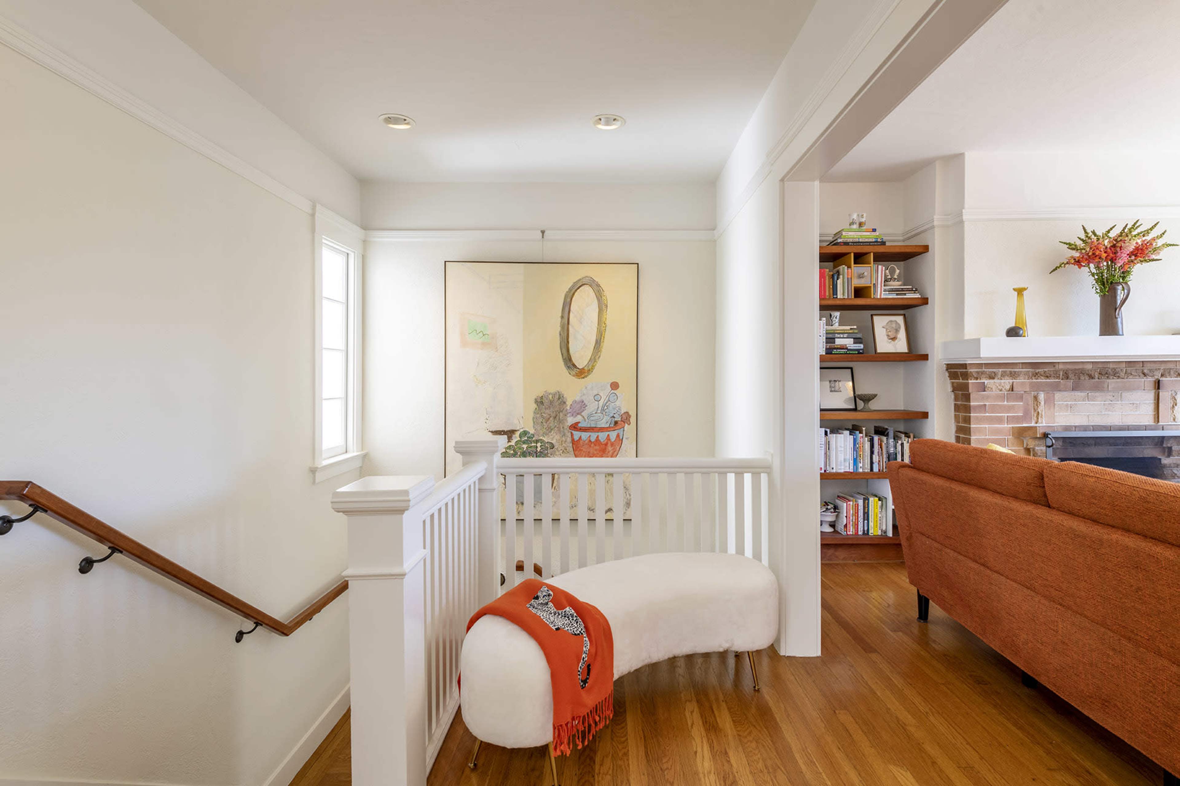 A cozy landing area with a curved white bench, a large painting on the wall, and a bookshelf alongside a living room featuring an orange couch.