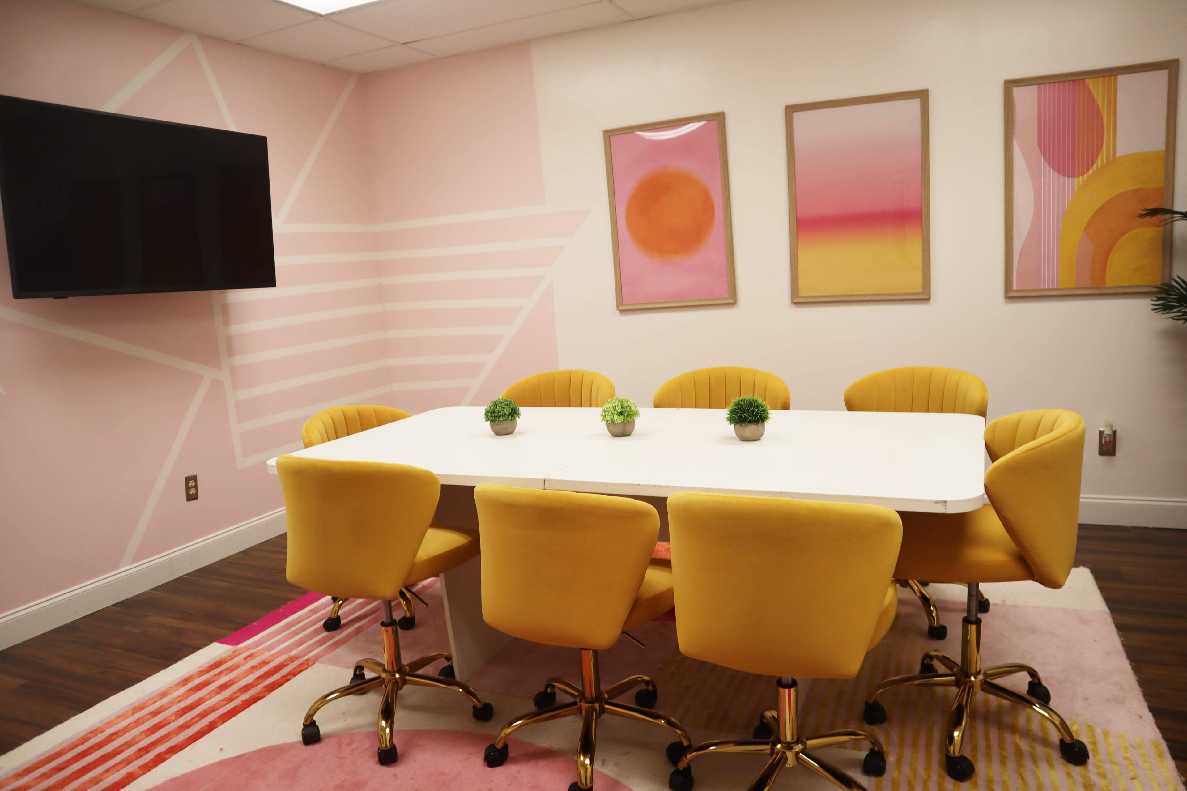A bright meeting room features a large white table surrounded by six yellow chairs and two framed artworks on the pink walls.