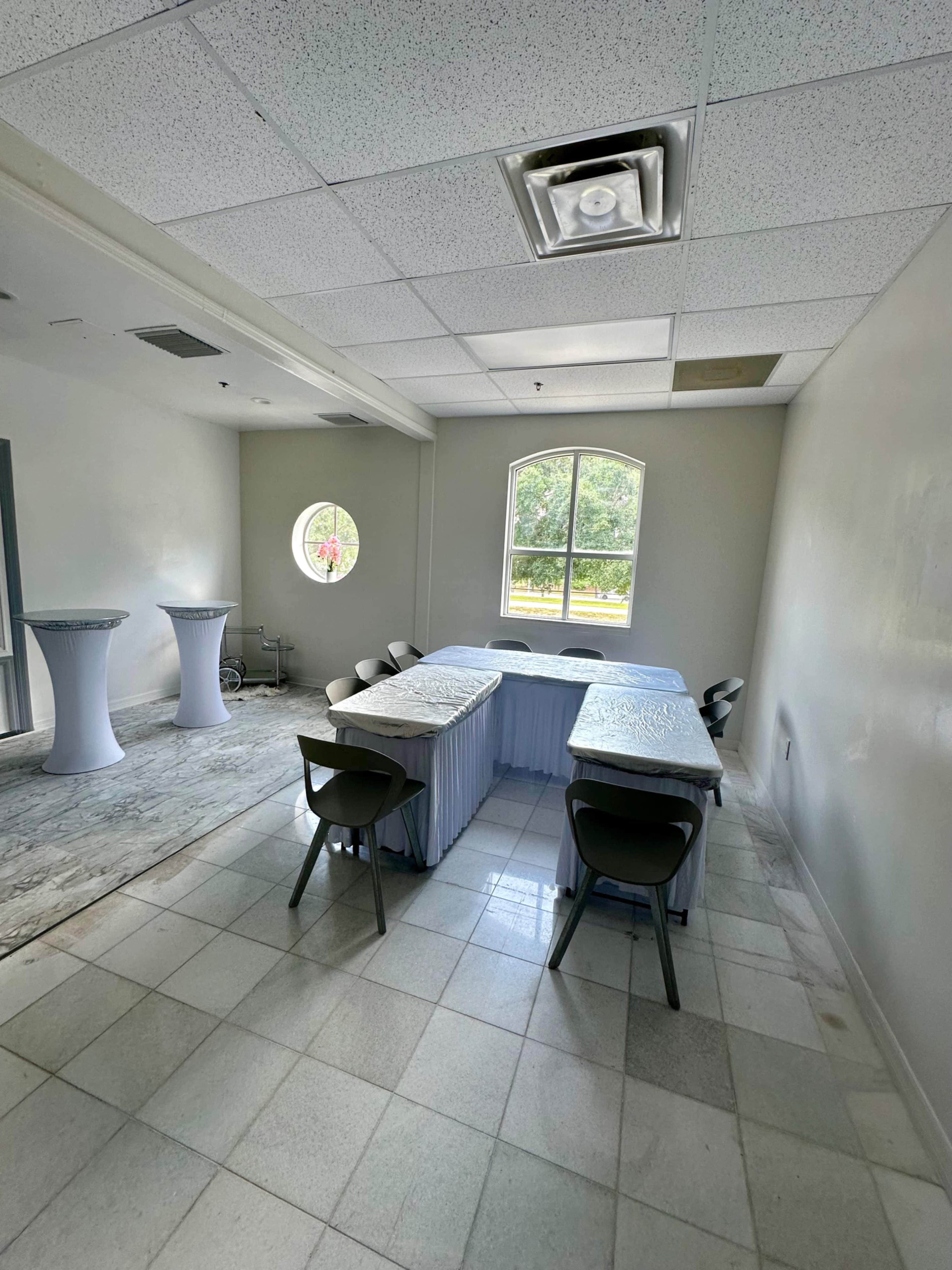 A small room with several tables arranged in a U-shape, surrounded by chairs, and featuring a circular window on one wall.