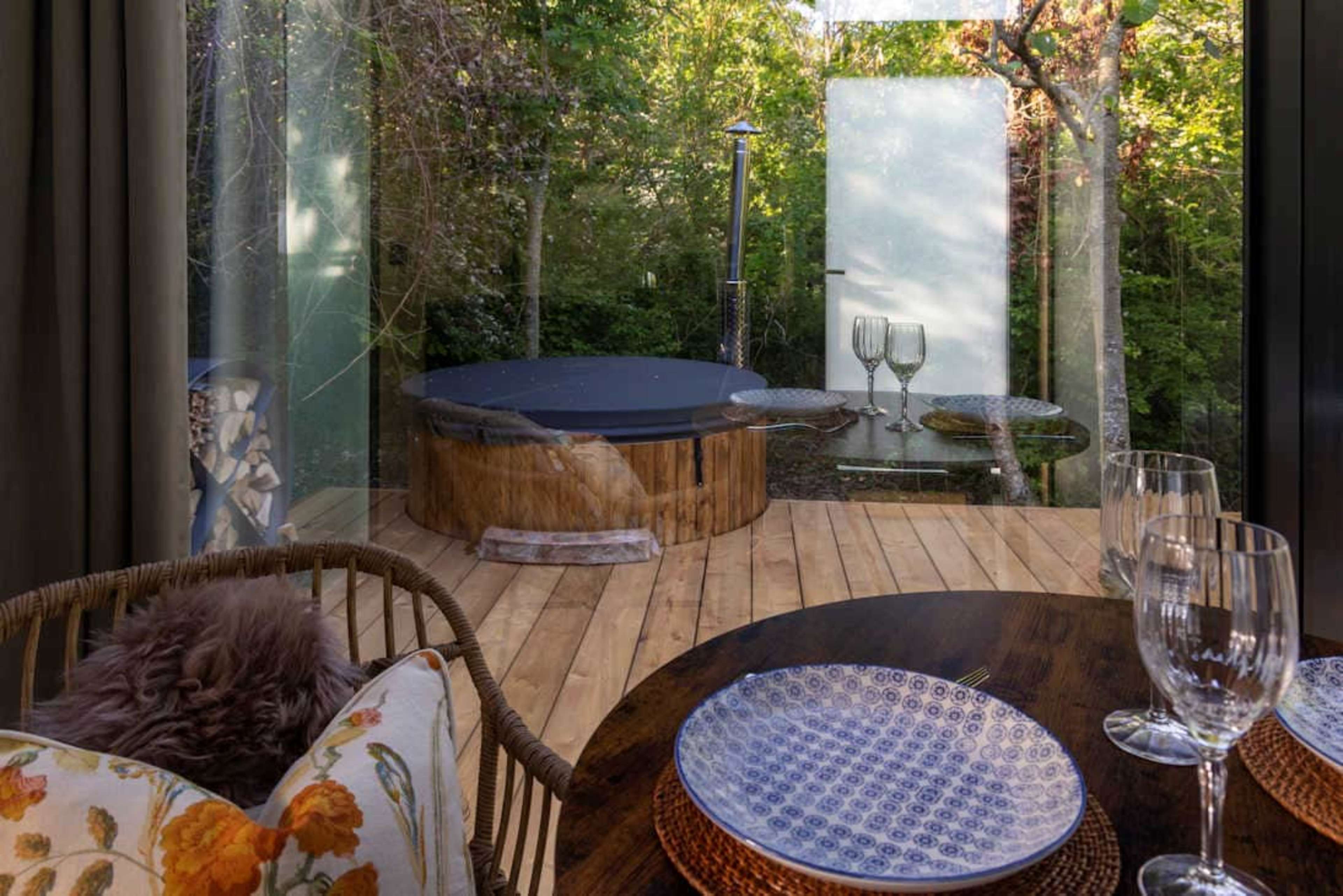 A modern dining area with wooden flooring, a round table set with plates and glasses, and a hot tub visible through a large glass window surrounded by greenery.