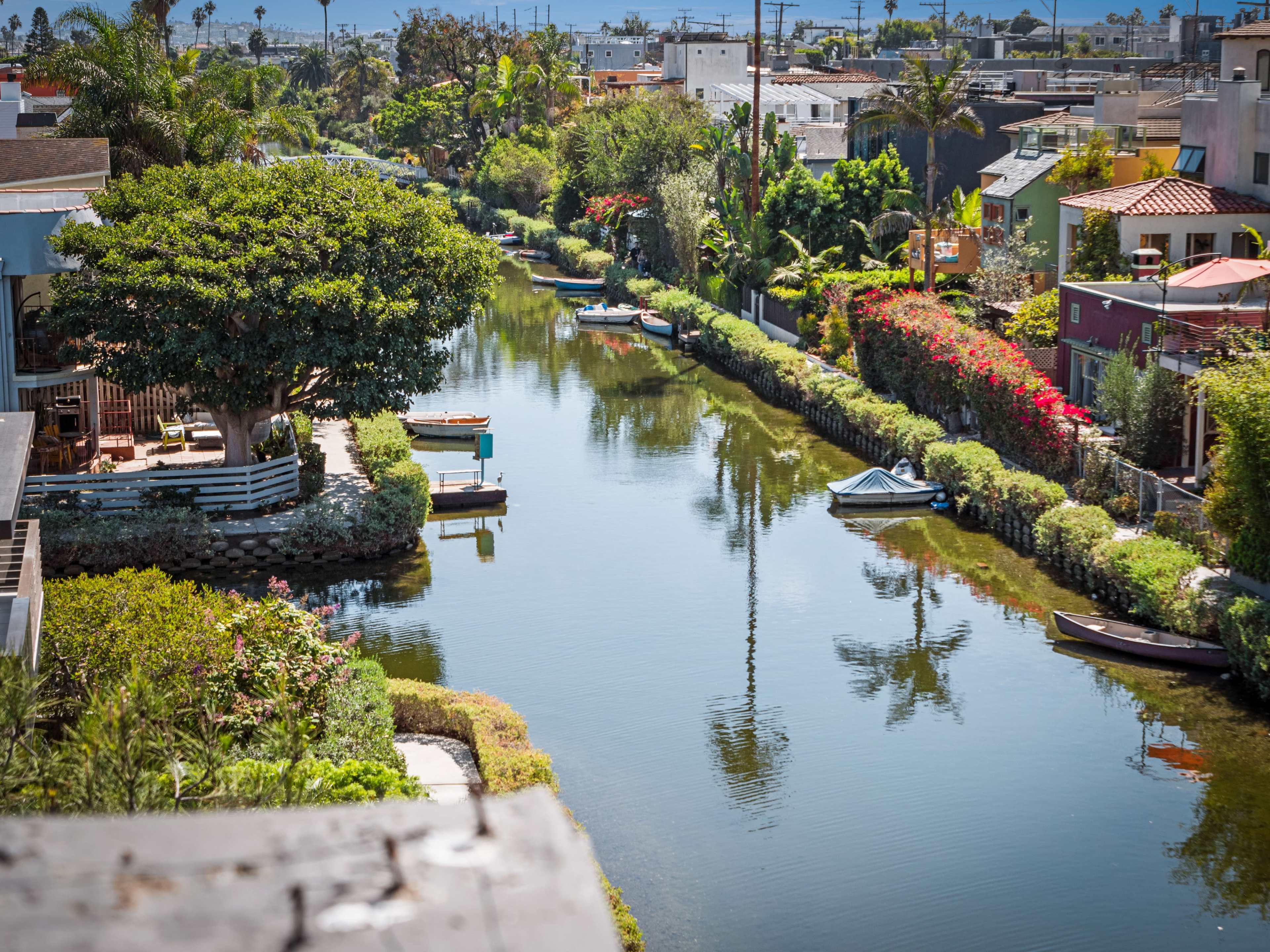 The image shows a peaceful canal lined with greenery and vibrant flowers, with small boats moored along the water's edge and houses visible in the background.