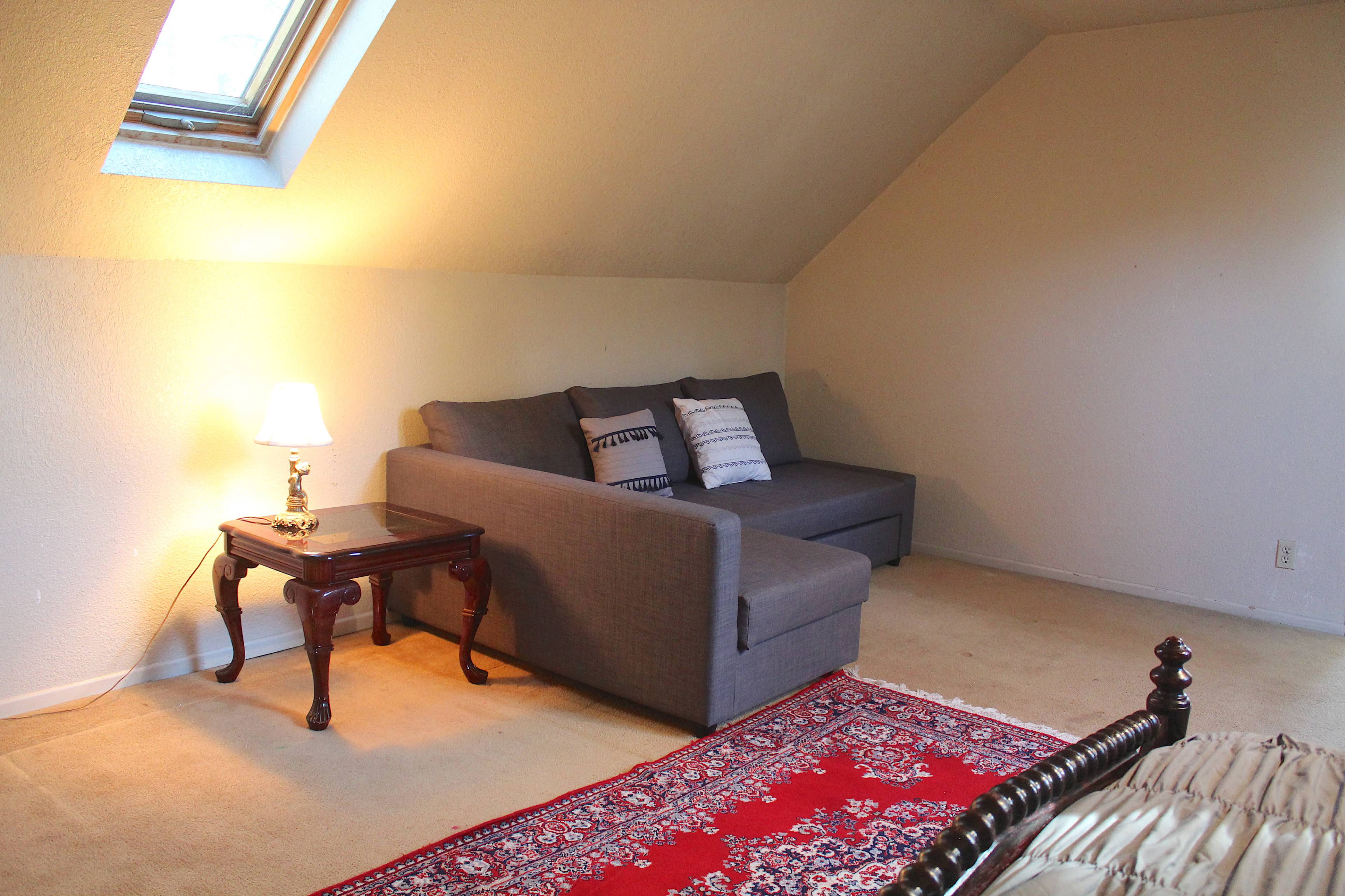 A cozy room with a sloped ceiling, featuring a gray couch, a small wooden side table with a lamp, and a decorative red area rug on beige carpet.