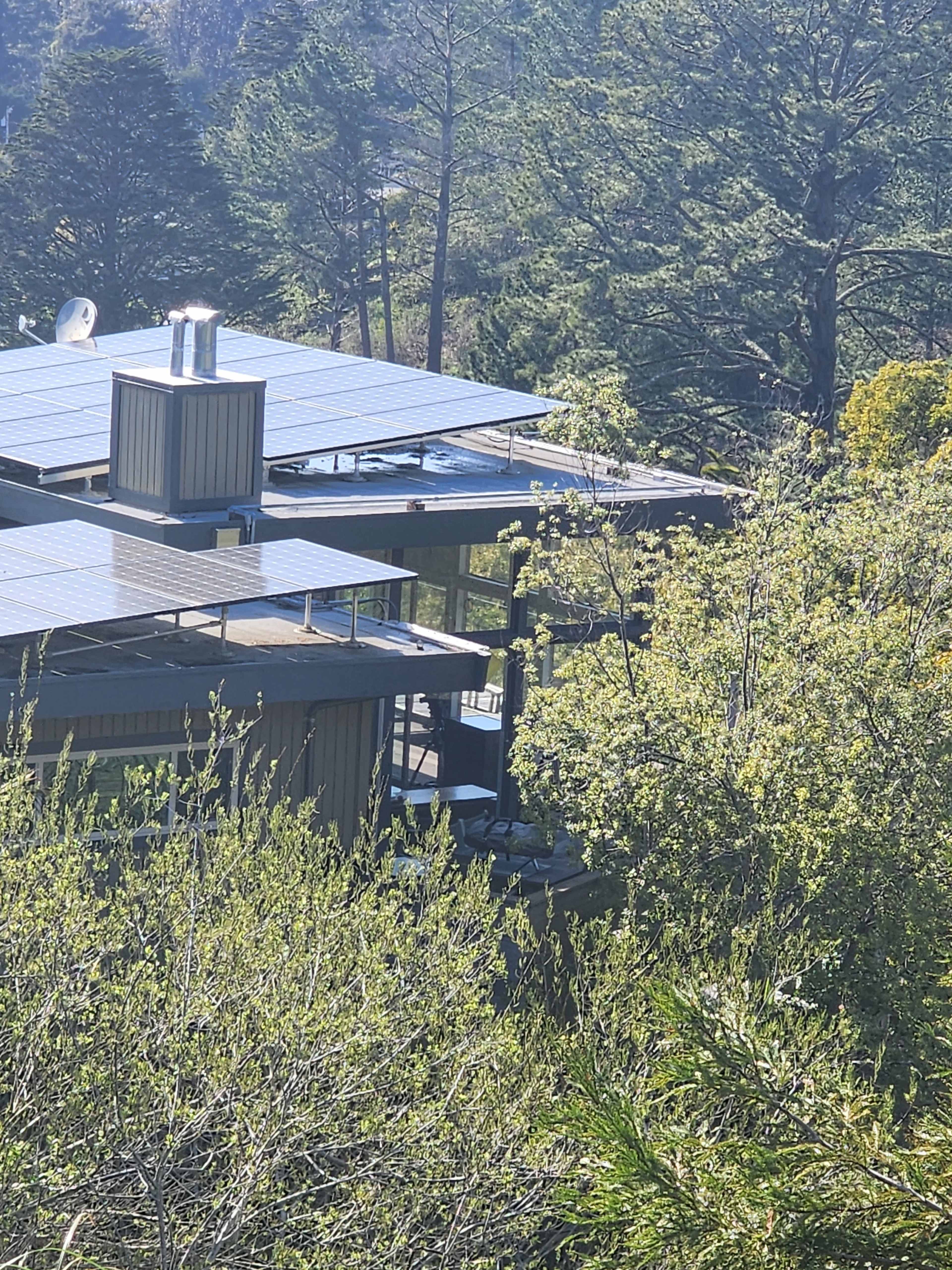 The image shows a modern house with solar panels on the roof, surrounded by trees and greenery.