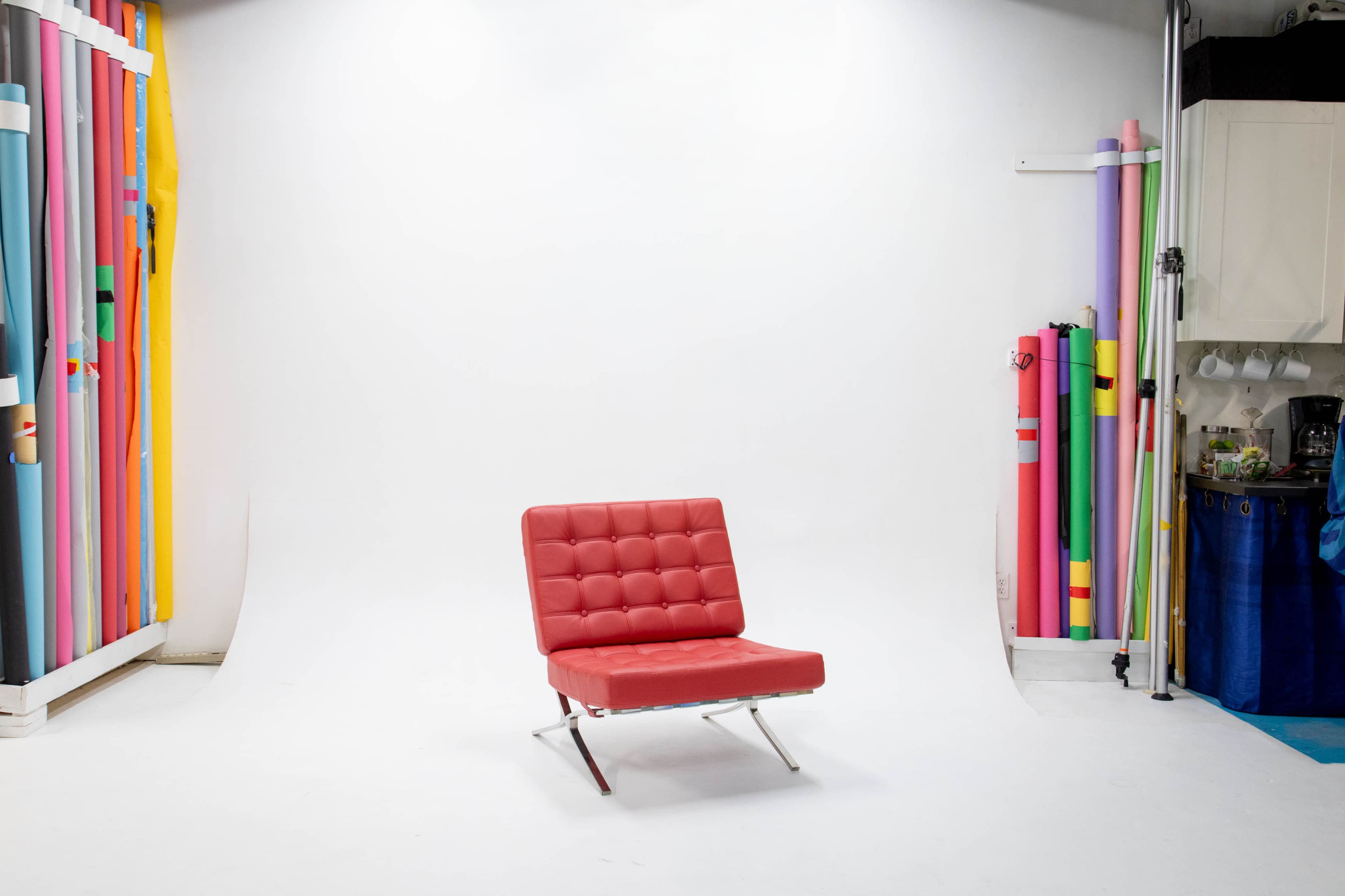 A red chair with a tufted design is positioned in the center of a photography studio with colorful backdrop rolls stacked to one side.