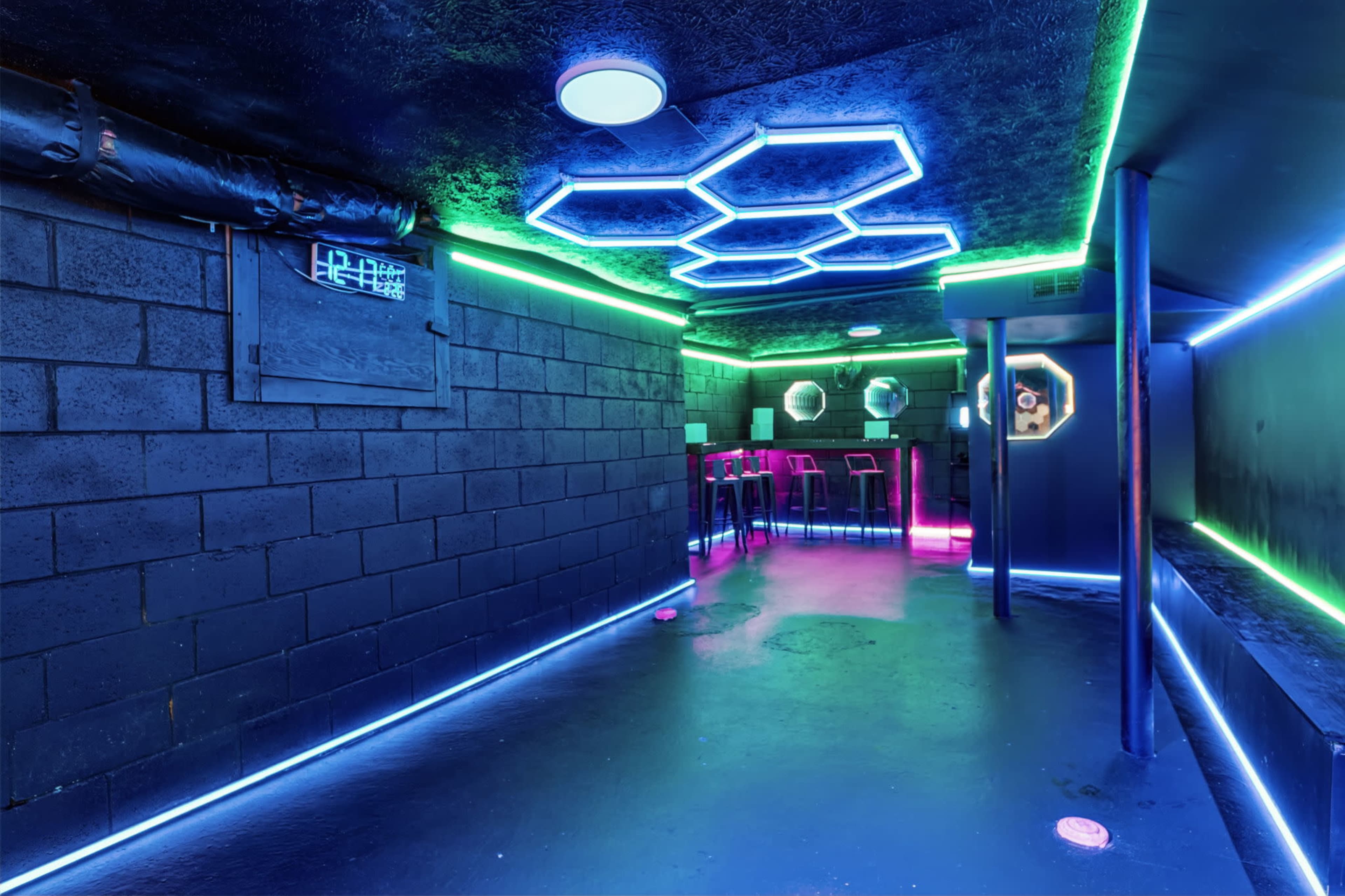 The image shows a dimly lit underground room with neon hexagonal lighting and a bar area featuring high stools against textured black walls.
