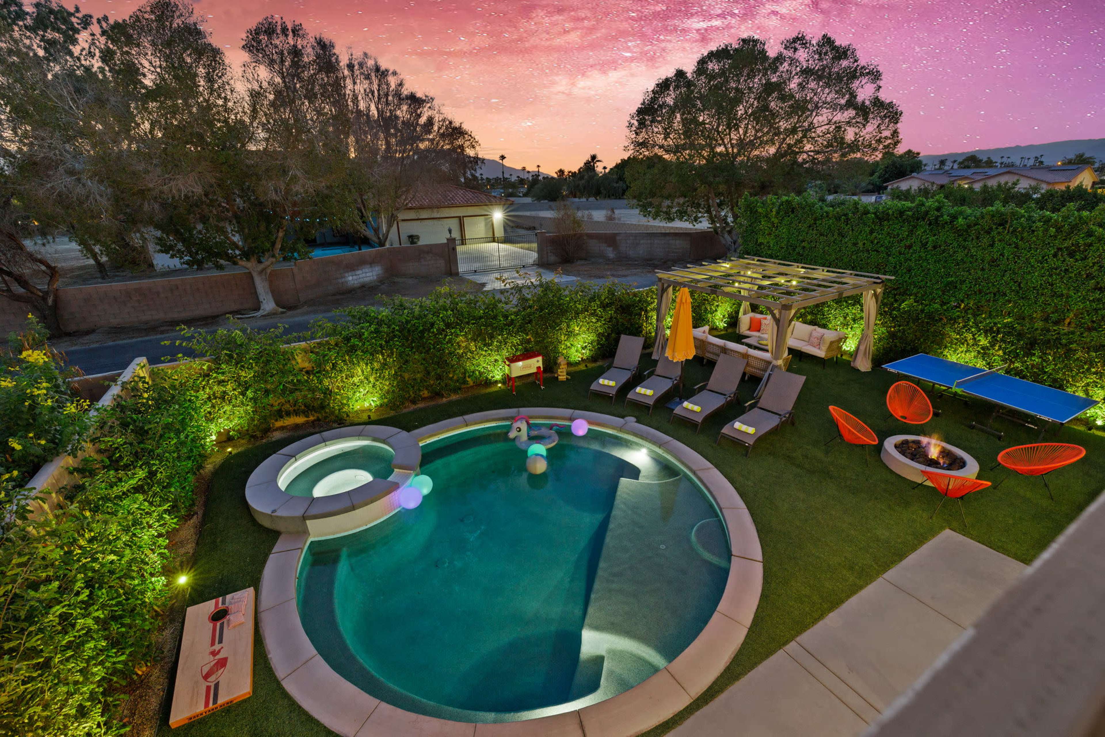 The image shows a backyard with a swimming pool surrounded by lounge chairs, a fire pit, and a covered seating area, all under a twilight sky.