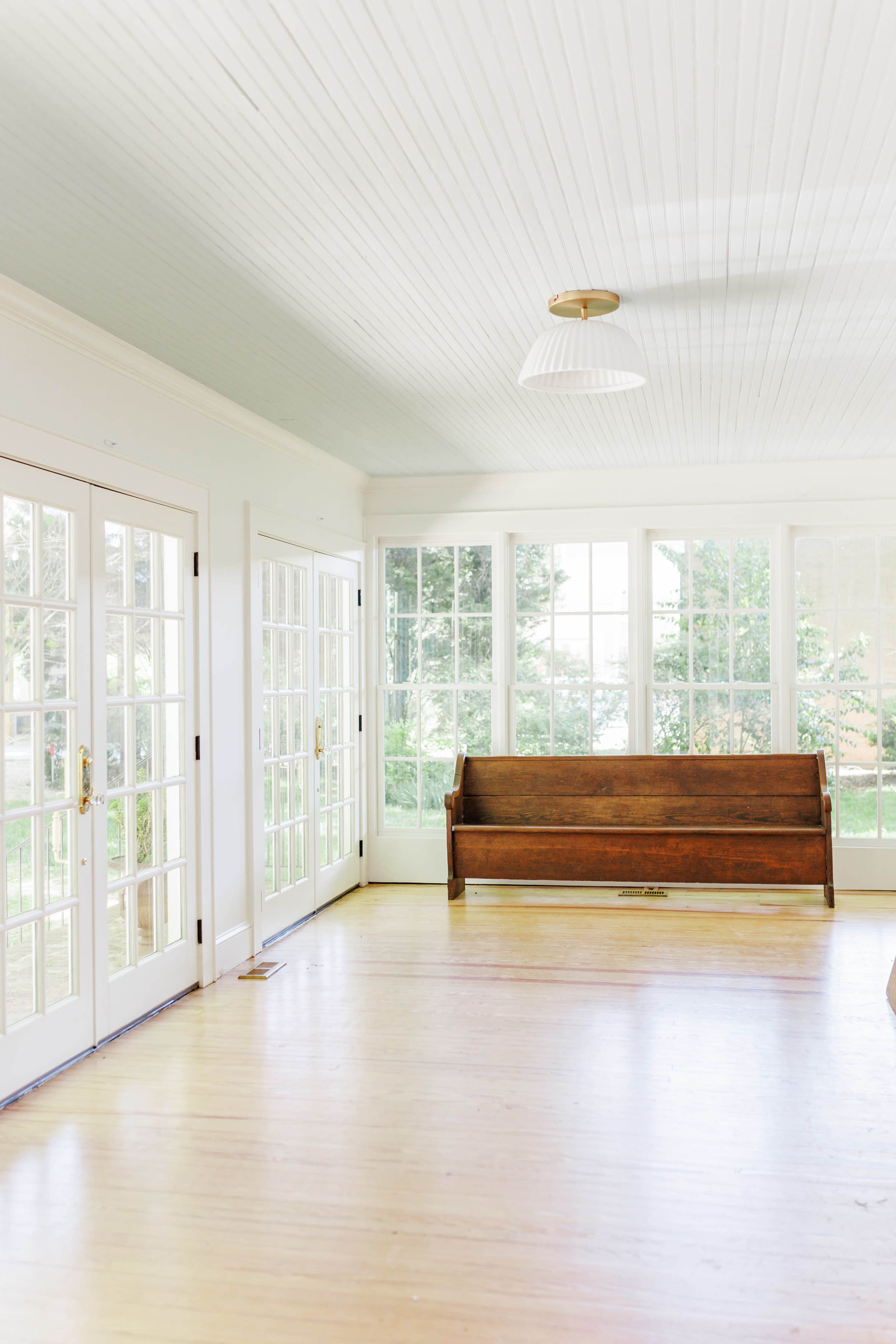 A wooden bench is positioned against a wall in a sunlit room featuring large windows and a wooden floor.