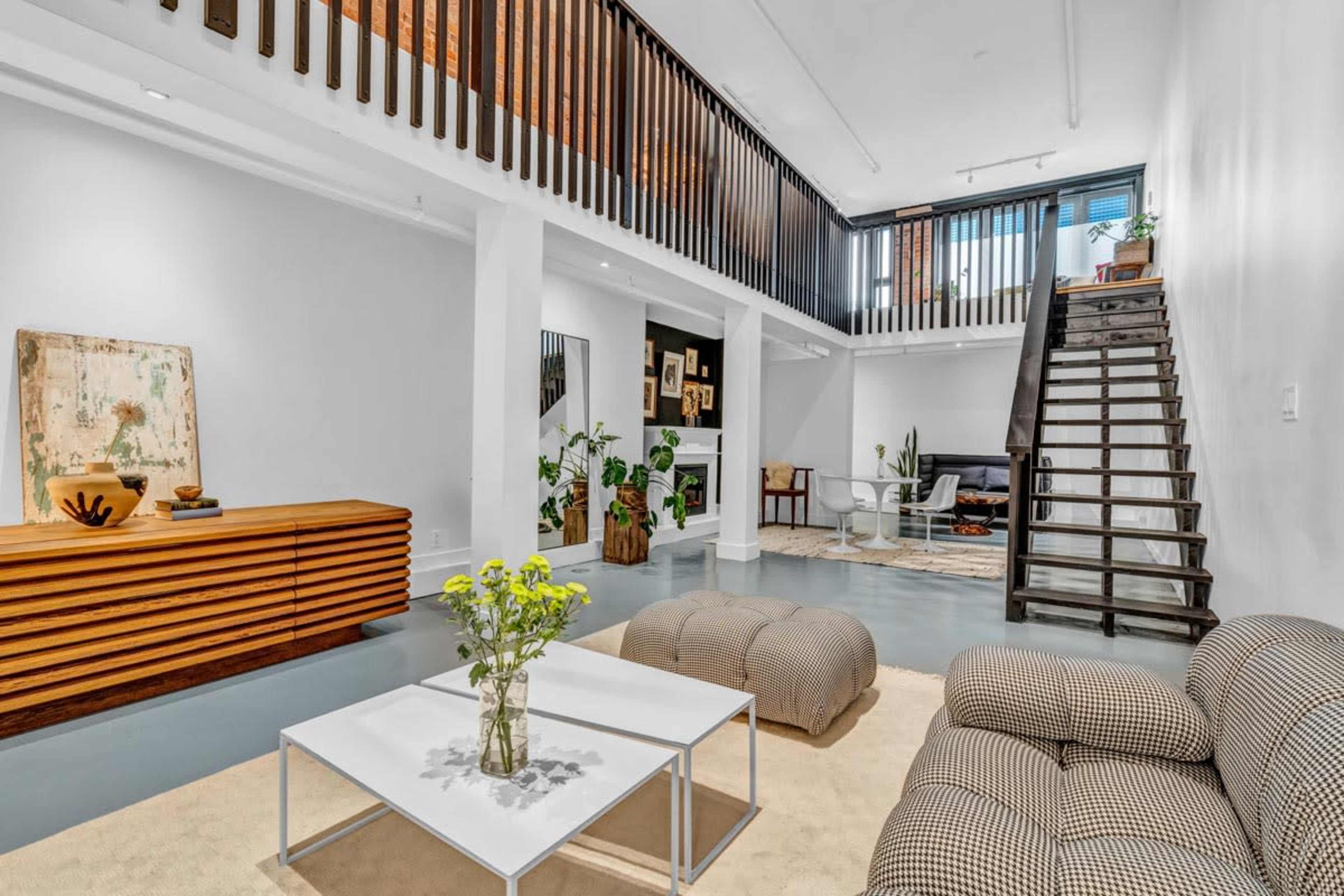 A spacious living area featuring a checkered sofa, a glass coffee table with a vase of flowers, and a staircase leading to a lofted space.